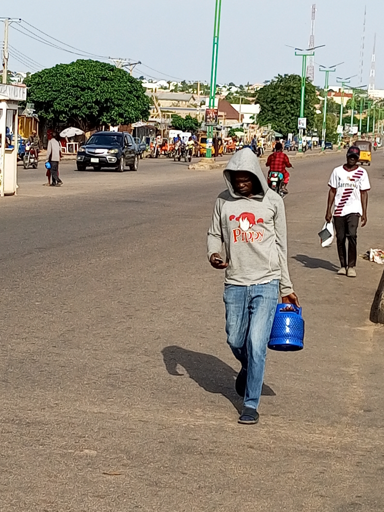 The image depicts a street scene in the city of Gombe, Nigeria. In the foreground, a man wearing a grey hooded sweatshirt with a "Pippy" logo and blue jeans walks towards the right of the frame, carrying a blue gas cylinder. He is also holding a mobile phone in his left hand. To his right, another man in a white and red striped shirt and black pants walks in the same direction, carrying a white bag. In the middle ground, a dark-colored SUV is driving on the road, and various people on motorcycles and a yellow motorized tricycle are visible. A large, leafy tree stands prominently behind the vehicles and people. The background shows buildings, more streetlights, and a hilly landscape. The overall impression is a busy urban environment with ongoing daily activities.