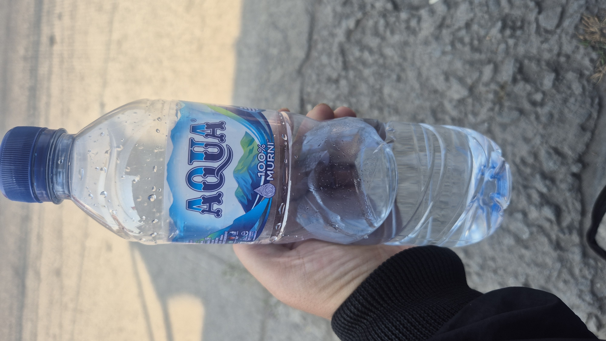 A person's hand, wearing a black jacket, is holding a clear plastic bottle of AQUA brand bottled water. The bottle is approximately half full, with condensation visible on the exterior. The label on the bottle features a blue and white design with mountains and the text "AQUA" and "100% MURNI". The background appears to be an outdoor setting with a sandy or dusty ground and a blurred shadow. The image is taken from a slightly low angle, focusing on the bottle and the hand holding it. The context of Karawang, Indonesia is not visually discernible from the image itself.