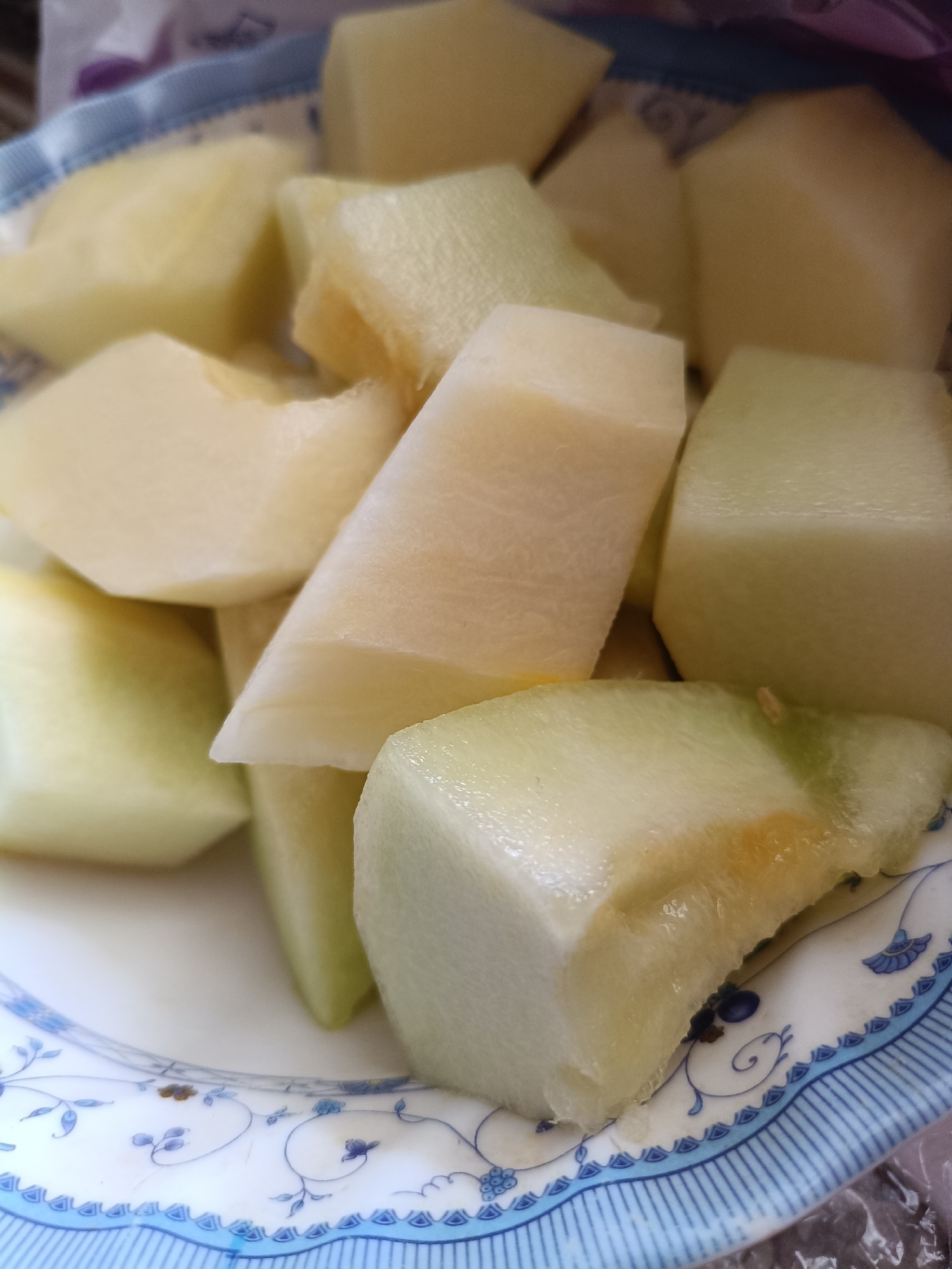 A close-up, top-down shot shows a white and blue patterned plate filled with chopped melon. The melon pieces are of various sizes and shapes, some are cubed, others are irregular chunks. The melon appears to be ripe, with a pale green rind visible on some pieces and a yellowish-white flesh. The plate has a decorative blue floral pattern along its rim. The background is out of focus but suggests an indoor setting, possibly a kitchen or dining area, with a hint of a plastic bag or wrapper visible at the top. There are no people or animals present in the image. The lighting is bright and seems to be coming from above, creating soft shadows on the fruit. The image does not contain any information that would allow for the determination of its location in Martuni, Armenia.