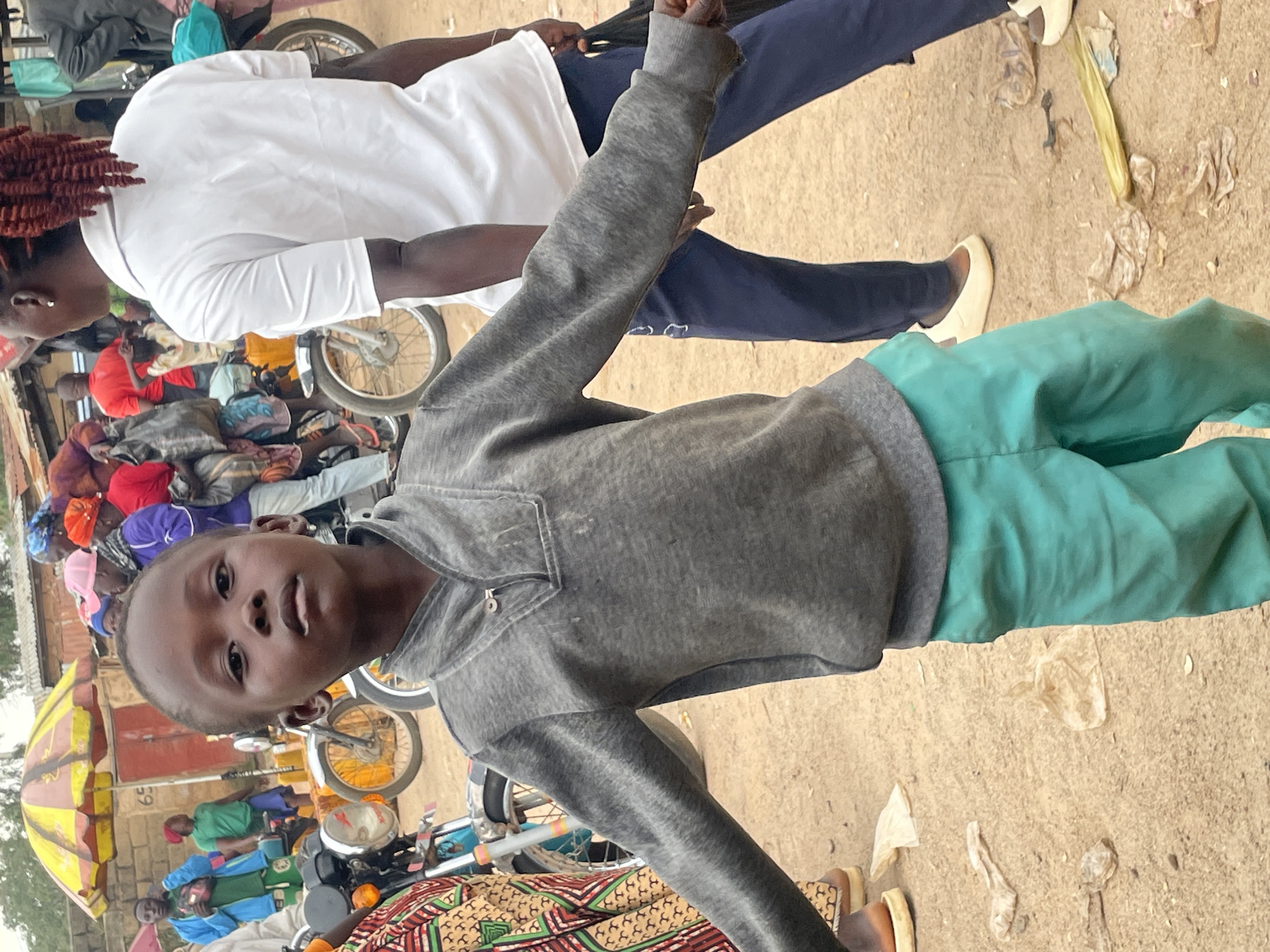 A young boy in a grey long-sleeved shirt and teal pants stands in a dusty outdoor market. He is looking directly at the camera with a serious expression. His arms are outstretched to his sides, and his hands are slightly open. Behind him, a woman in a white polo shirt and dark blue pants stands with her back to the camera. Other people and motorcycles are visible in the background, along with a yellow and red umbrella and buildings. The ground is covered in dirt and some scattered debris, including pieces of plastic. This scene appears to be in a busy marketplace in Wang, Nigeria.