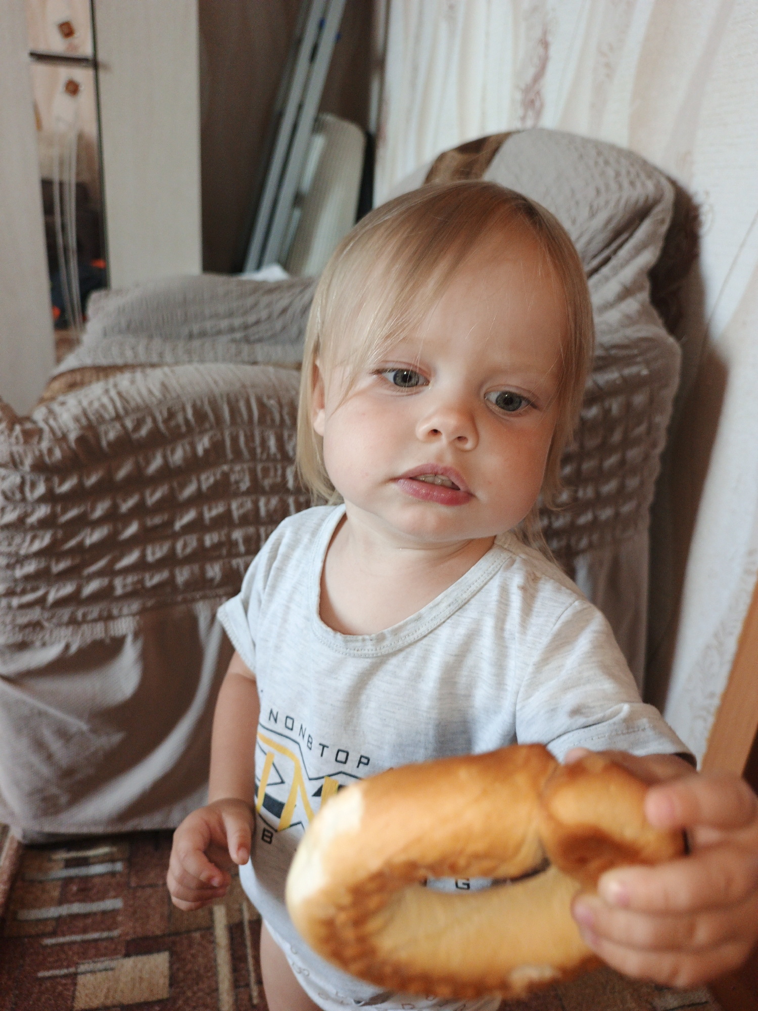 The image shows a close-up shot of a toddler with blonde hair and blue eyes, holding a large ring-shaped pastry in their hands. The child is wearing a light gray t-shirt with a graphic design. The background suggests an indoor setting, possibly a home, with furniture covered in a patterned fabric and curtains. The overall lighting is natural, indicating it may be daytime. The scene does not provide any information to place it in Medvedyovka, Russia, or any other specific city.