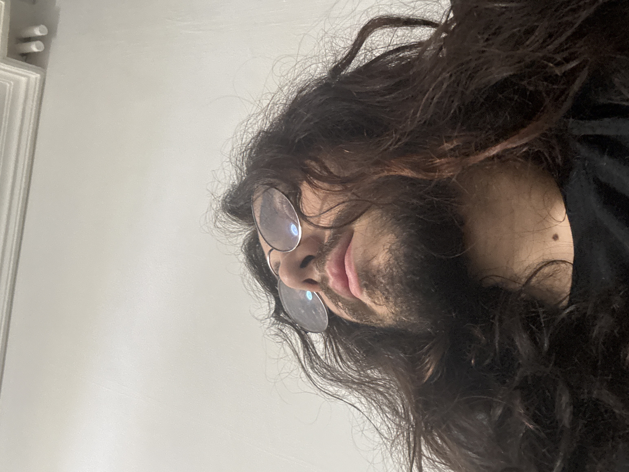 A close-up, low-angle shot shows the face of a person with long, dark, messy hair and a beard, looking upwards. The person is wearing round eyeglasses with reflective lenses. Their lips are slightly parted. The background is a plain, off-white wall with molding at the top. The image appears to have been taken indoors in an urban environment, likely within a residential or commercial building. The lighting is even and diffused, suggesting ambient light or indirect artificial lighting. The scene is static, with no apparent action or interaction occurring. The focus is primarily on the individual's face and hair, with the background providing context.