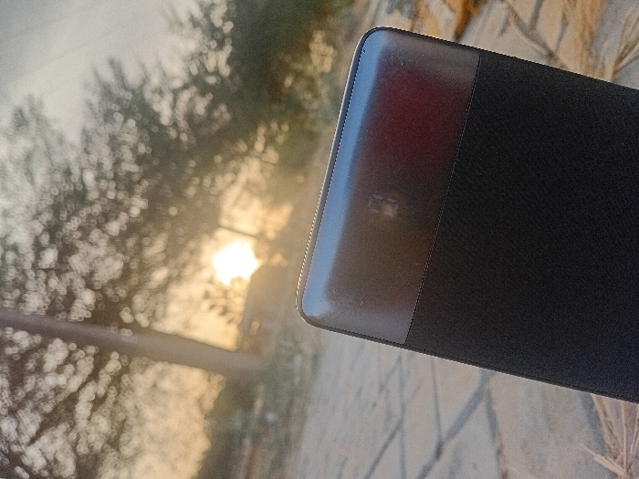 A dark, rectangular object with rounded edges dominates the right side of the frame, possibly a book or tablet case. It features a shiny, reddish-brown upper section contrasting with a matte black surface below. The background is blurred, revealing trees and a bright, diffused light source, likely the sun. The scene appears to be outdoors, possibly during sunrise or sunset, and a textured surface is partially visible beneath the object.