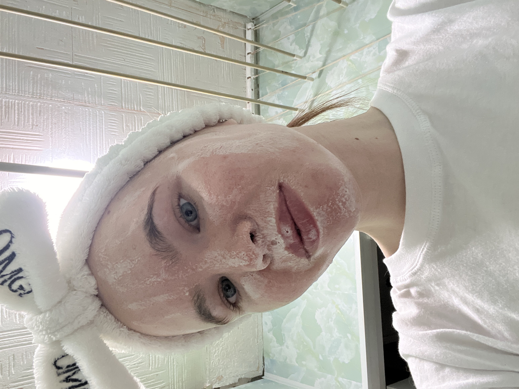 A woman with blue eyes and fair skin is depicted close-up, facing the camera at an angle. She has a white fluffy headband with the letters "OMG" on one of the ears, and her face appears to have a white cosmetic substance applied to it.
The background is somewhat indistinct, but reveals a light green tiled wall pattern and a white textured surface, possibly a ceiling or wall. A metal clothes drying rack is suspended overhead. The context suggests this is likely a bathroom setting in Chaikovsky, Russia.