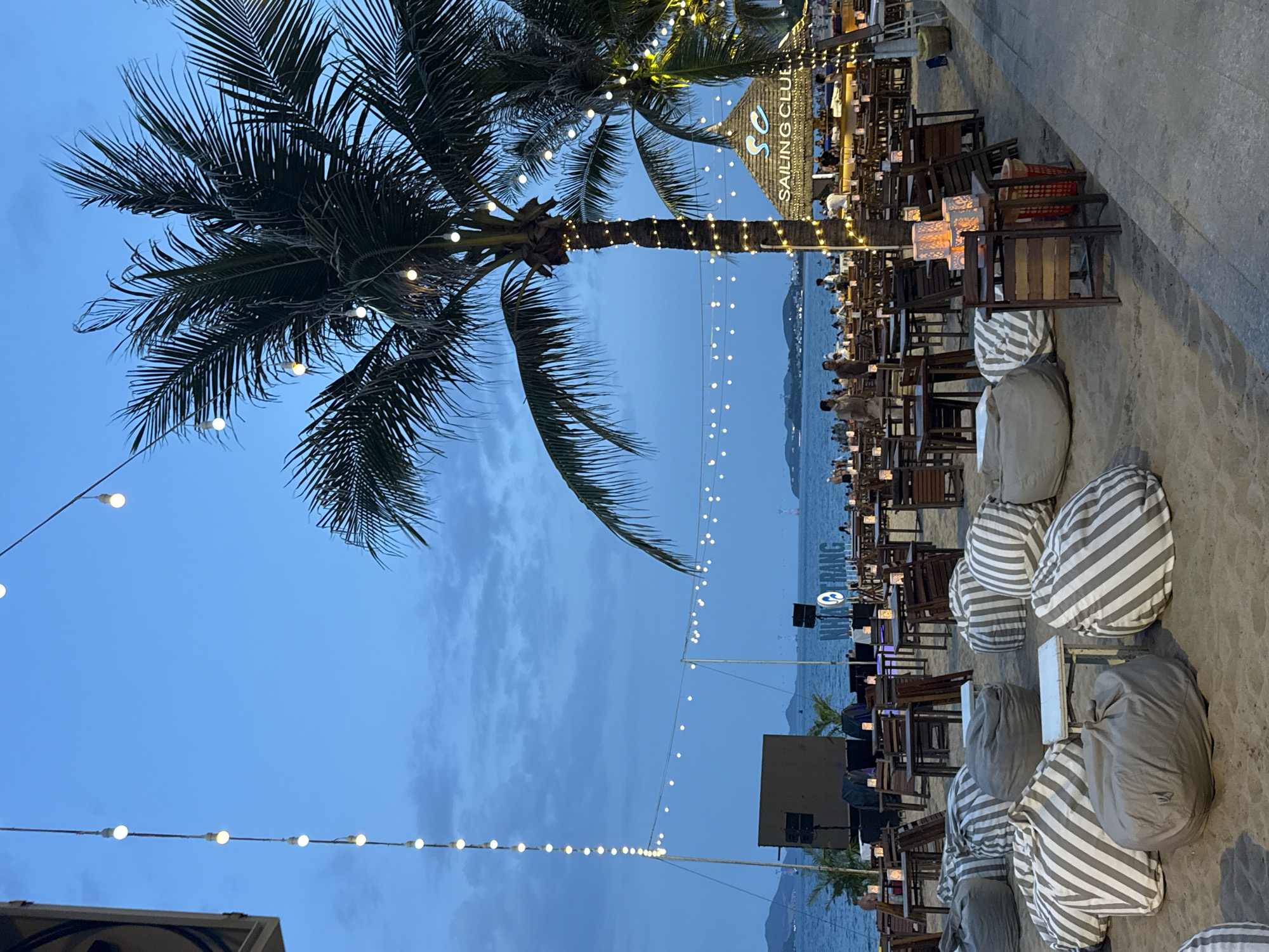 The image shows an outdoor evening scene at the Sailing Club in Nha Trang, Vietnam. Tables and chairs, along with bean bag chairs, are set up on a beach sand surface, illuminated by string lights and decorative lanterns. A palm tree decorated with lights stands to the left. In the background, the sea is visible, and distant mountains or landmasses are silhouetted against the evening sky. Patrons are seated at some of the tables. The "Sailing Club" sign is visible above the tables.