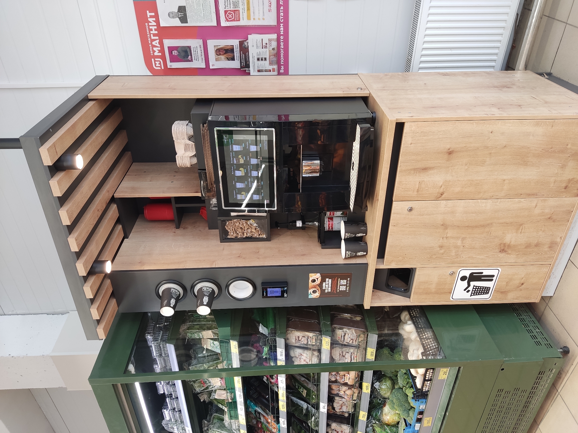 A vending machine is visible in a commercial setting, possibly a store, with vegetables and food items inside. The machine includes a coffee maker, cups, and a trash receptacle. The vending machine is likely located in Krasnodar, Russia, as indicated by the text in Russian on the sign above the machine. The food in the vending machine includes various vegetables and mushrooms. There is a spotlight on the upper left side of the vending machine. Prices are marked on the vegetable items.