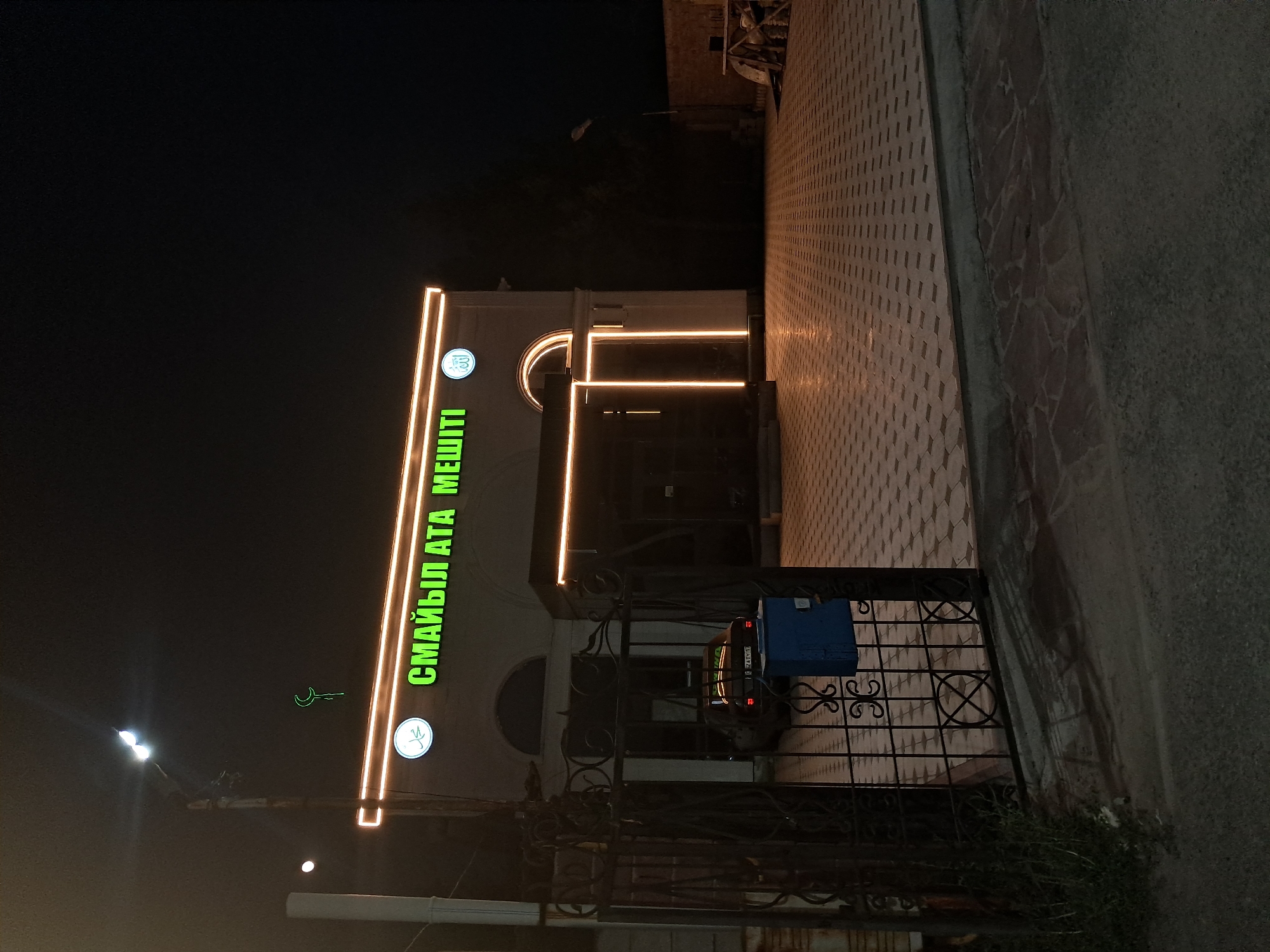 This is a night-time photograph taken outdoors, possibly in Shymkent, Kazakhstan. The main subject is a building with illuminated signage. The building appears to be a mosque, as indicated by the Arabic script on the sign and a crescent moon motif. The sign, written in green neon letters, reads "СМАЙЫЛ АТА МЕШІТІ" which translates to "Smail Ata Mosque".
The building has a modern architectural style with large windows and glowing orange neon accents along its facade, outlining geometric shapes and architectural features. In front of the building, there is a decorative metal fence and a small blue mailbox. A portion of a vehicle is visible behind the fence.
The ground in front of the building is paved with a patterned tile, and to the right of the paved area is a rougher, textured surface that appears to be a road or sidewalk. The overall atmosphere is dark due to the time of day, with artificial lighting from the building and a street lamp creating a stark contrast. The weather is not discernible from the image. The scene appears quiet and still, suggesting a typical evening outside a place of worship.