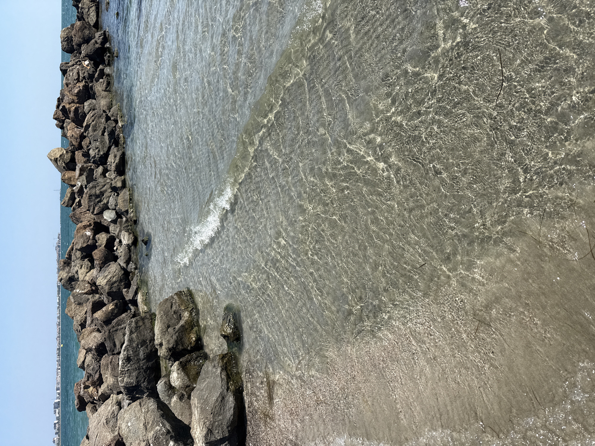 This image shows a clear, shallow sea with a rocky breakwater extending into the water. The water is calm, with small waves lapping at the sandy shore. The seabed is visible through the clear water, showing a mixture of sand and small stones. In the background, a city skyline can be seen on the horizon, with buildings and a radio tower. The sky is clear and blue. The scene appears to be located on the coast of Pomorie, Bulgaria, as suggested by the presence of the city skyline and the characteristics of the coastline. There are no people visible in the image.