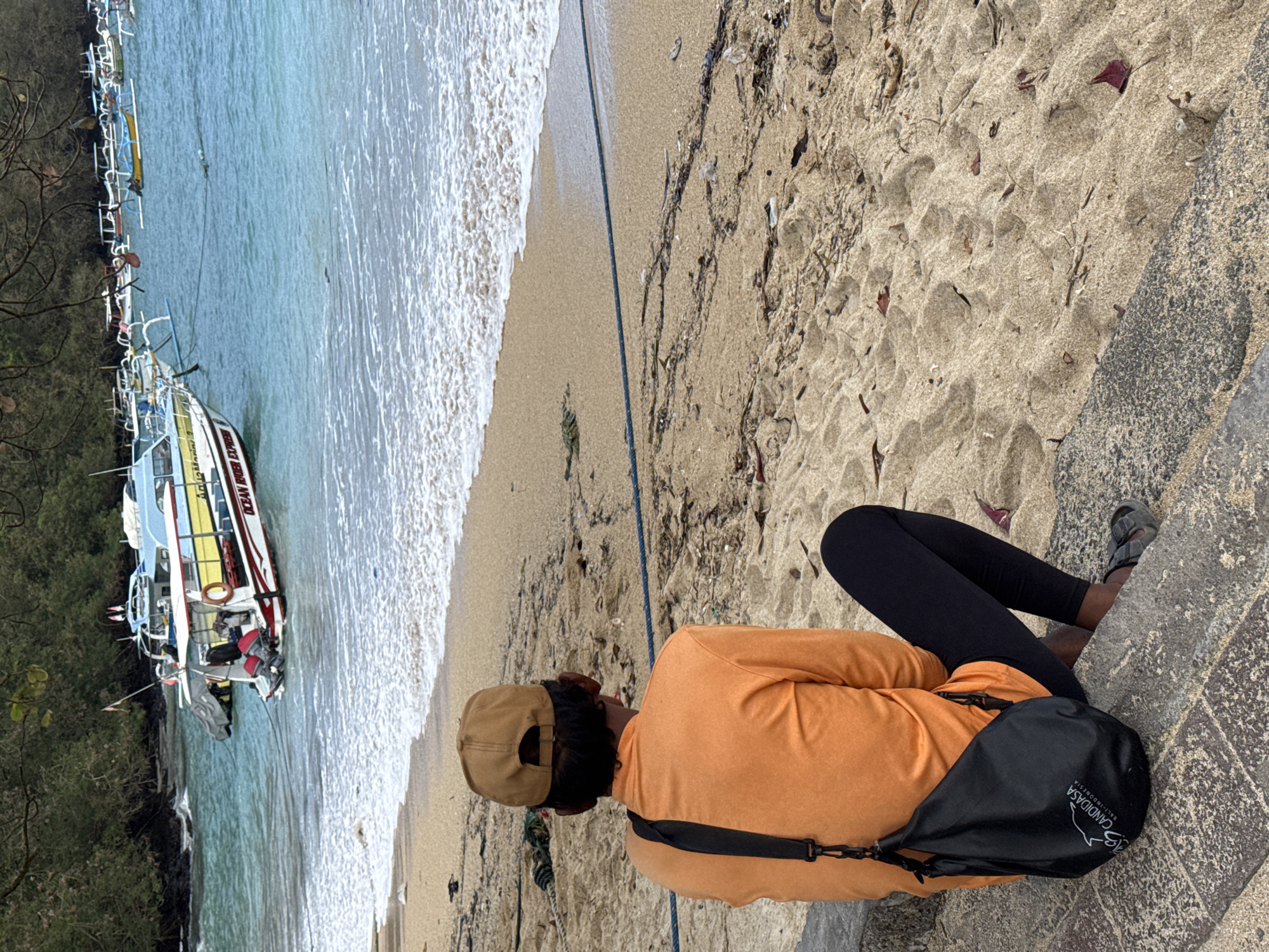 An over-the-shoulder medium shot captures a person from behind, seated on concrete steps leading down to a sandy beach. The person wears a brown baseball cap, an orange long-sleeved shirt, black leggings, and dark sandals. A black crossbody bag with a white logo reading "CANDIDASA BALI-INDONESIA" is slung over their shoulder. The beach is strewn with debris including leaves, twigs, and plastic. Gentle waves break on the shore. In the background, the turquoise ocean is dotted with numerous traditional Balinese fishing boats and several larger speedboats. The water is calm, and the sky is overcast, suggesting a potentially humid day. The scene is set in Candidasa, a coastal town in the Manggis district of Bali, Indonesia, known for its serene beaches and traditional boat culture.