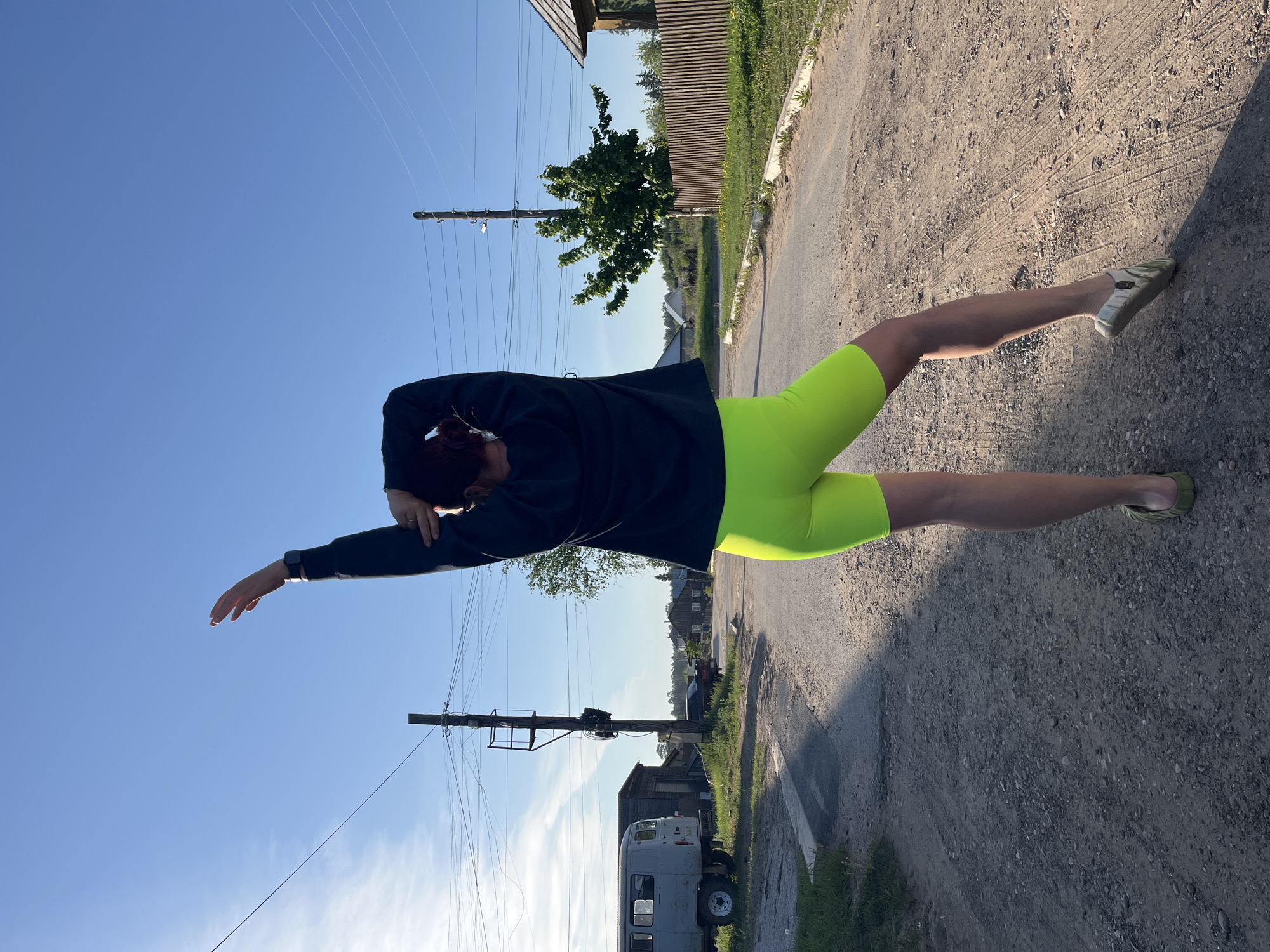 A photograph captured in what appears to be a rural or semi-rural area of Chaikovsky, Russia, depicts a person standing on a dirt road in an unorthodox pose. The individual is oriented almost horizontally in the frame, wearing a dark, long-sleeved shirt and bright neon-yellow cycling shorts. One arm is extended towards the clear, blue sky, while the other arm is bent with the hand near the head.
The surroundings consist of a worn dirt road with patches of asphalt. To the right, the road surface gives way to a shadow. Buildings are visible in the background, including a wooden structure and a light-colored van or truck parked alongside the road. Power lines extend across the sky. A green tree is visible, providing a backdrop to the scene.