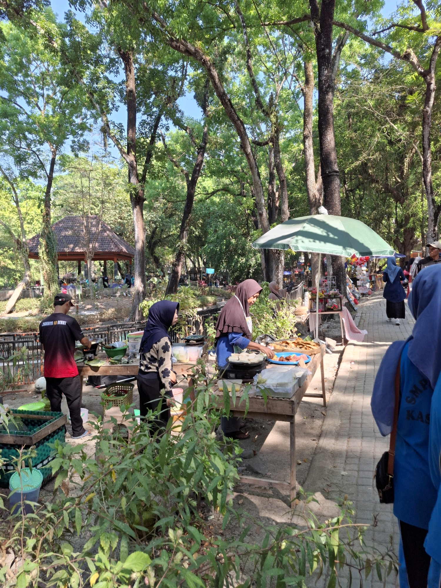 In Tawun, Indonesia, a scene unfolds in a park-like setting under a canopy of lush green trees. The ground is a mix of dirt and a paved walkway, with a small body of water or drainage visible to the left. Several food stalls are set up, with one woman in a brown hijab and blue shirt actively cooking on a portable stove, preparing food on a metal tray. Another person, wearing a black shirt with red accents and a cap, stands near a stove, also engaged in food preparation. A woman in a black hijab and patterned shirt is observing the cooking. A large green umbrella provides shade over a stall displaying various goods, including colorful flowers and trinkets. Further down the walkway, other individuals are present, including a person in a blue hijab and another in a blue shirt with "SMK KAS..." visible on the back. Baskets and containers filled with produce and food items are scattered around the stalls. In the background, a traditional Javanese gazebo structure with a brown tiled roof is visible, suggesting a recreational area within the park. The overall atmosphere appears to be a bustling outdoor market or food gathering place.
