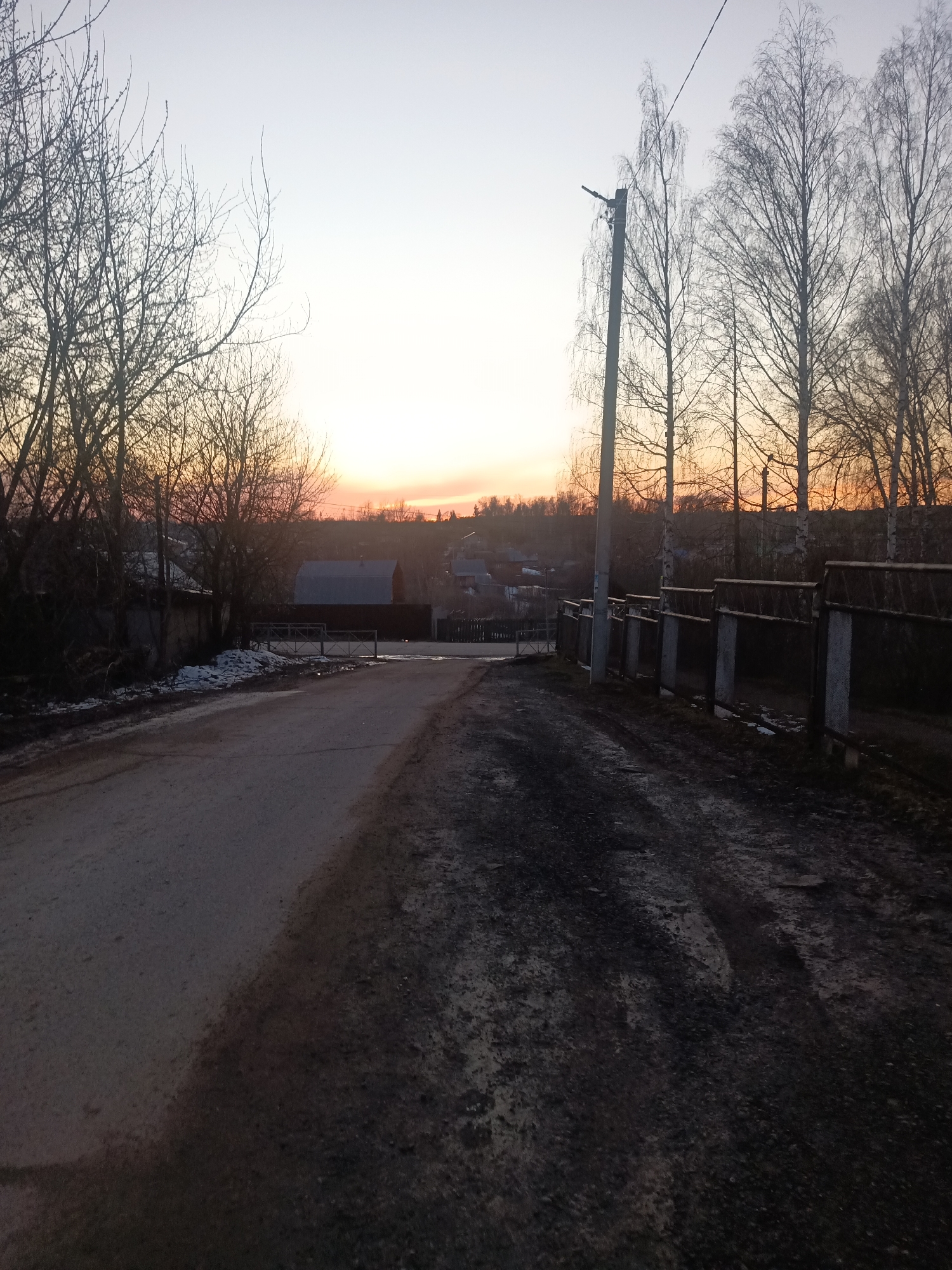 The image captures a street scene in Фоки, Russia, at dusk. The road, partially paved, is divided into a smoother, lighter gray section on the left and a darker, rougher, dirtier section on the right. The sky is a gradient of pale hues, transitioning from a bright, almost white, horizon line to a light blue-gray above. Bare trees line both sides of the road, their branches silhouetted against the twilight sky. A weathered wooden fence runs along the right side of the road, supported by concrete posts. In the background, glimpses of houses and buildings can be seen, indicating a residential area. A utility pole stands on the right side of the road, near the fence. The ground is uneven and appears muddy in places. Overall, the scene conveys a sense of quiet rural or suburban ambiance during the transition from day to night.