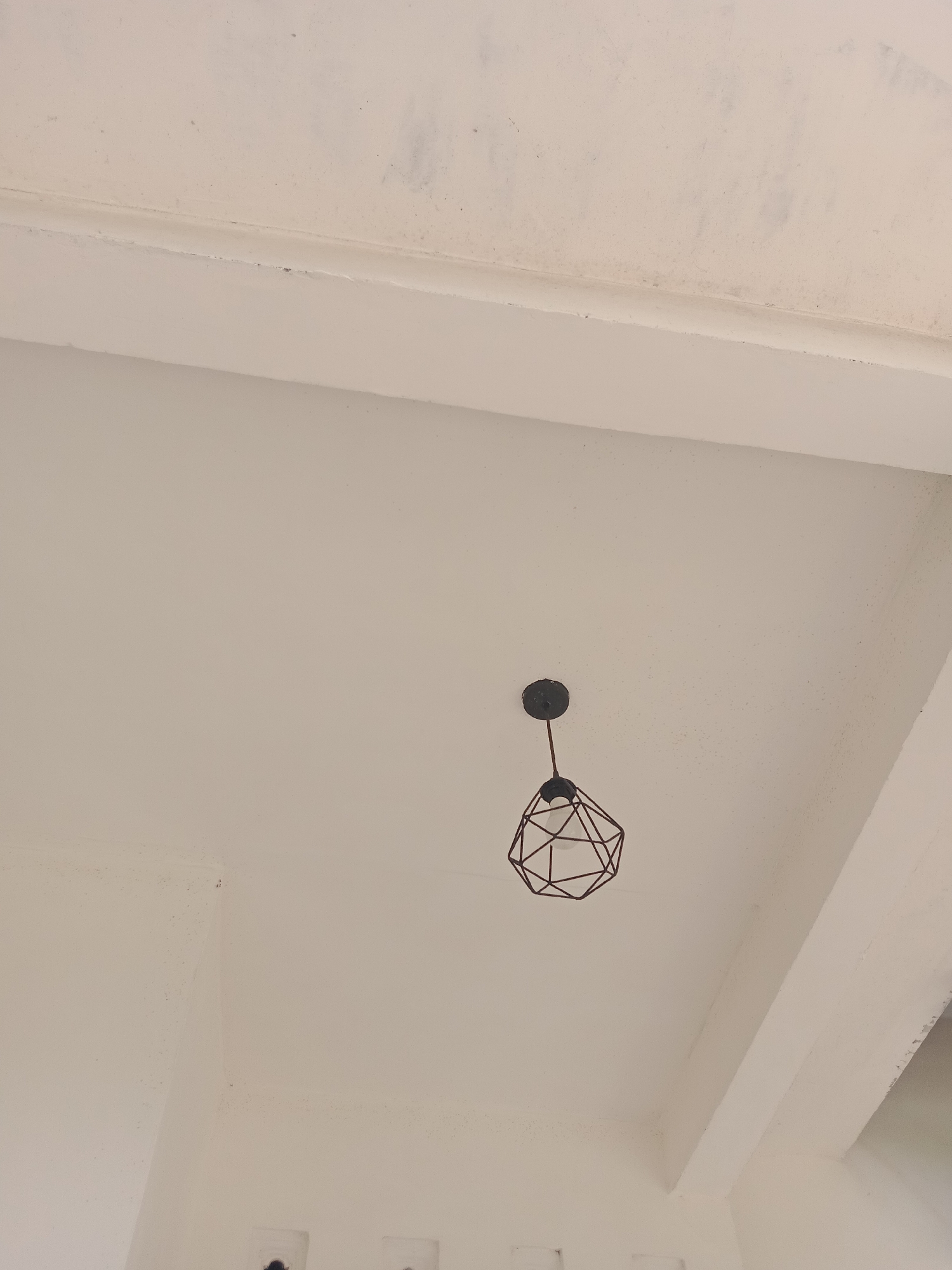 The image shows a close-up view of a white ceiling with a dark geometric pendant light fixture hanging from it. The fixture has a cage-like design. There are some minor imperfections and dark spots on the ceiling, suggesting it may be textured or slightly aged. The lighting in the room appears to be coming from the fixture and possibly an unseen source. The perspective suggests the photo was taken from a slightly low angle, looking up at the ceiling. There are no people or active events depicted. The location is identified as Stiaji, Indonesia.
