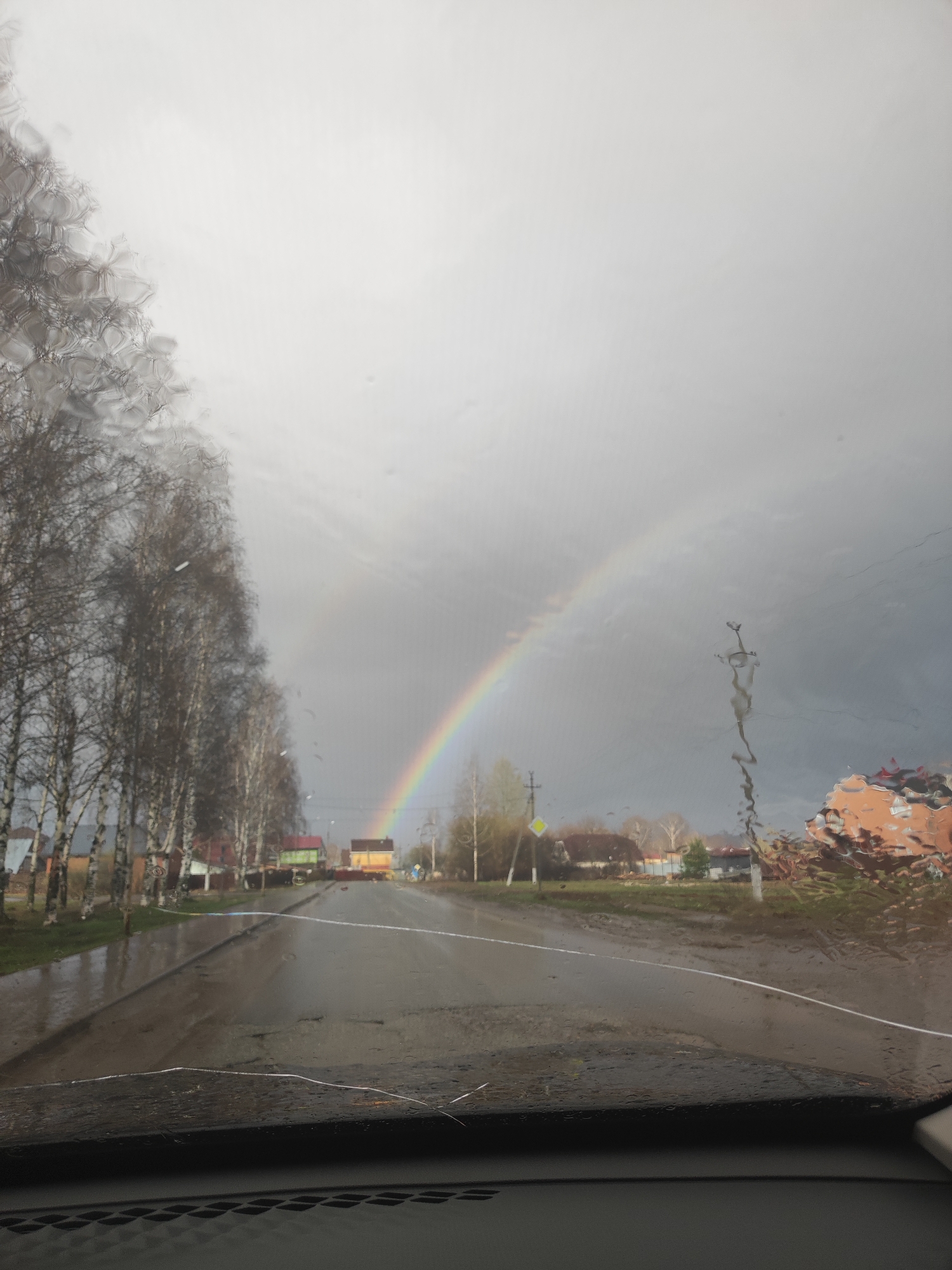 The image shows a view from inside a car looking out onto a street after a rain shower. A rainbow is visible in the distance. The road is wet and reflective, and a row of bare trees lines the left side of the street. Houses are visible in the distance on both sides of the street. The foreground includes the car's dashboard and a partially obscured view through the windshield, which has water droplets on it. This image was taken in the city of Votkinsk, Russia.