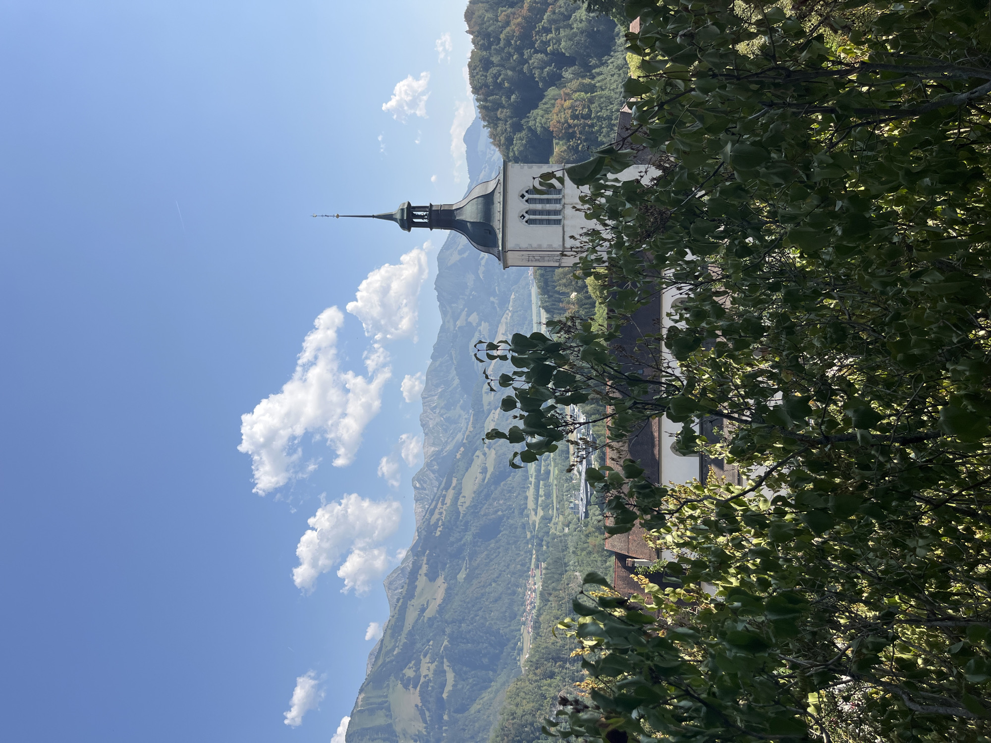 The image shows a view of a church tower and surrounding landscape, likely in Epagny, Switzerland. The church tower is white with a dark, steeple-like roof and arched windows. In front of the tower, there are trees with green leaves, partially obscuring the view. Behind the church, there are rolling hills covered in lush green trees, with a small town nestled in the valley. Further in the distance, majestic mountains rise, their peaks possibly snow-capped, under a bright blue sky dotted with fluffy white clouds. The overall impression is one of a serene, picturesque rural setting.