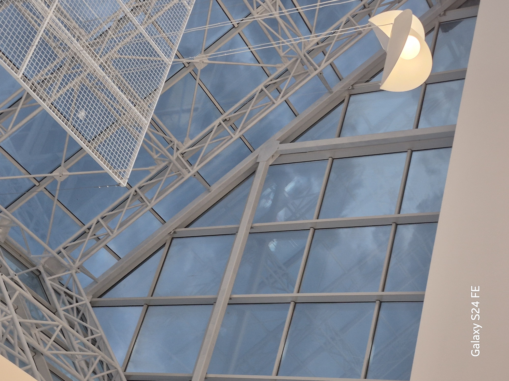 The image shows an interior view of a modern building's ceiling and upper walls, likely a conservatory or a space with a large glass roof. The structure features a white geometric framework supporting large glass panels that reflect a cloudy blue sky. A prominent, contemporary white pendant light fixture hangs from the ceiling. In the bottom left corner, the text "Galaxy S24 FE" is visible, indicating the device used to capture the image. There are no people or animals present in the scene. The overall impression is one of architectural design and natural light. There is no information within the image to suggest it is located in Chișinău, Moldova.