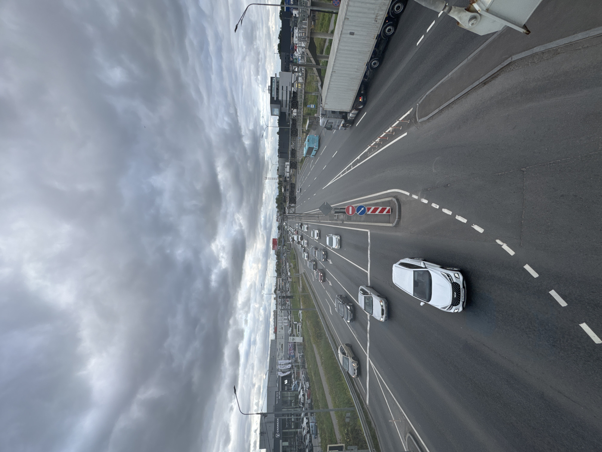 The image shows a highway scene under a cloudy sky in Saint Petersburg, Russia. Several cars, including a white SUV in the foreground, are visible on the multi-lane highway. A large truck with a white container is on the right side of the road. In the background, there are buildings, including a car dealership with "CHANGAN" signage, and streetlights. The road has white lane markings and is bordered by grass and barriers. The weather appears overcast, contributing to a muted color palette.