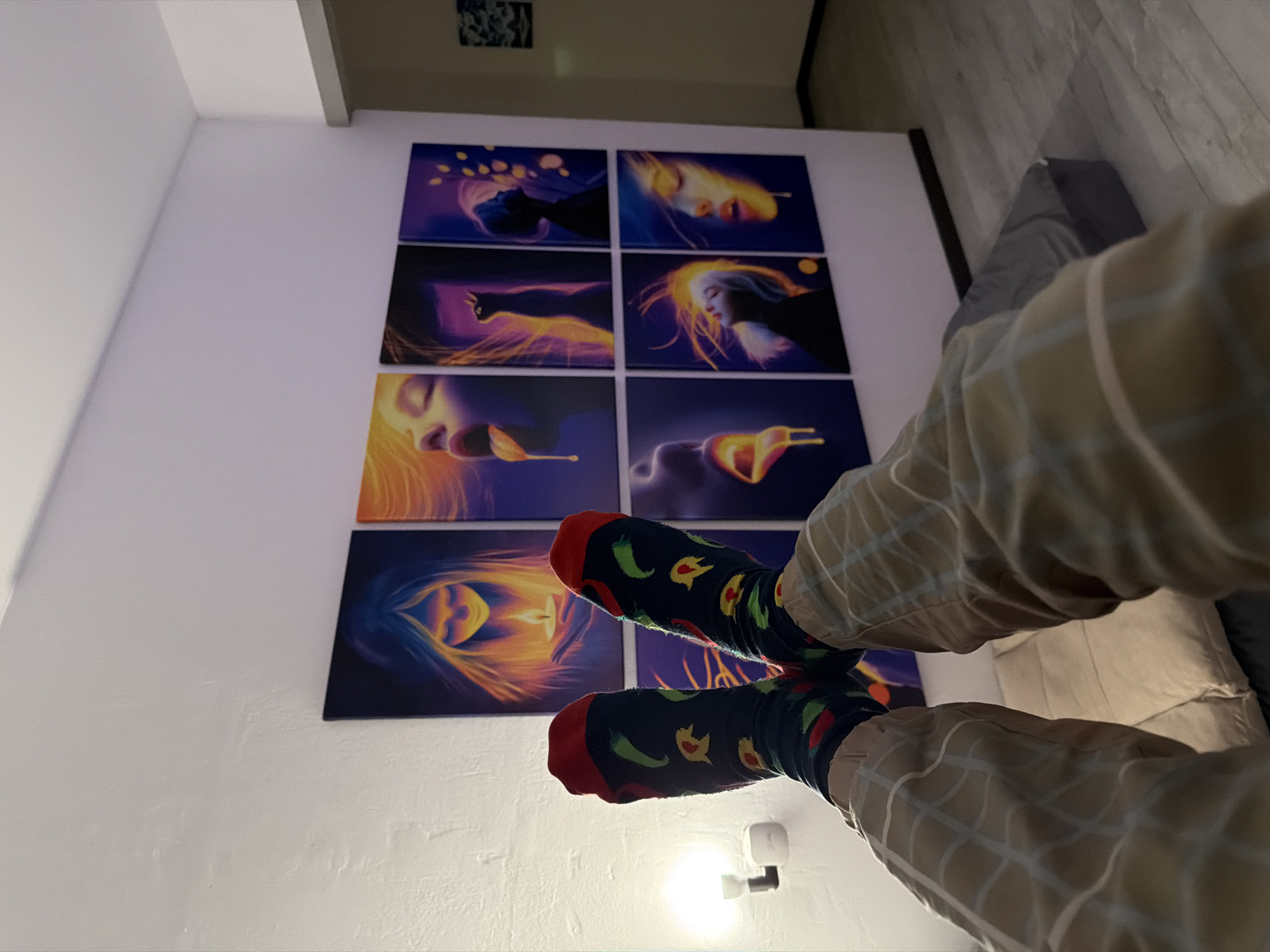 The image shows a person's legs and feet wearing patterned socks and plaid pants, resting in front of a wall adorned with a grid of eight colorful, abstract art pieces. The artwork is displayed on canvas and features stylized depictions of faces, a cat, and other abstract elements in vibrant colors. The room appears to be a living space, with a white wall, a light fixture on the left, and a floor with a wood-like finish. A dark grey cushion or mattress is visible on the floor in the lower right quadrant. The scene is lit by ambient light, creating a somewhat moody atmosphere. There is no specific indication of location within Naberezhnye Chelny, Russia, or any other city.
