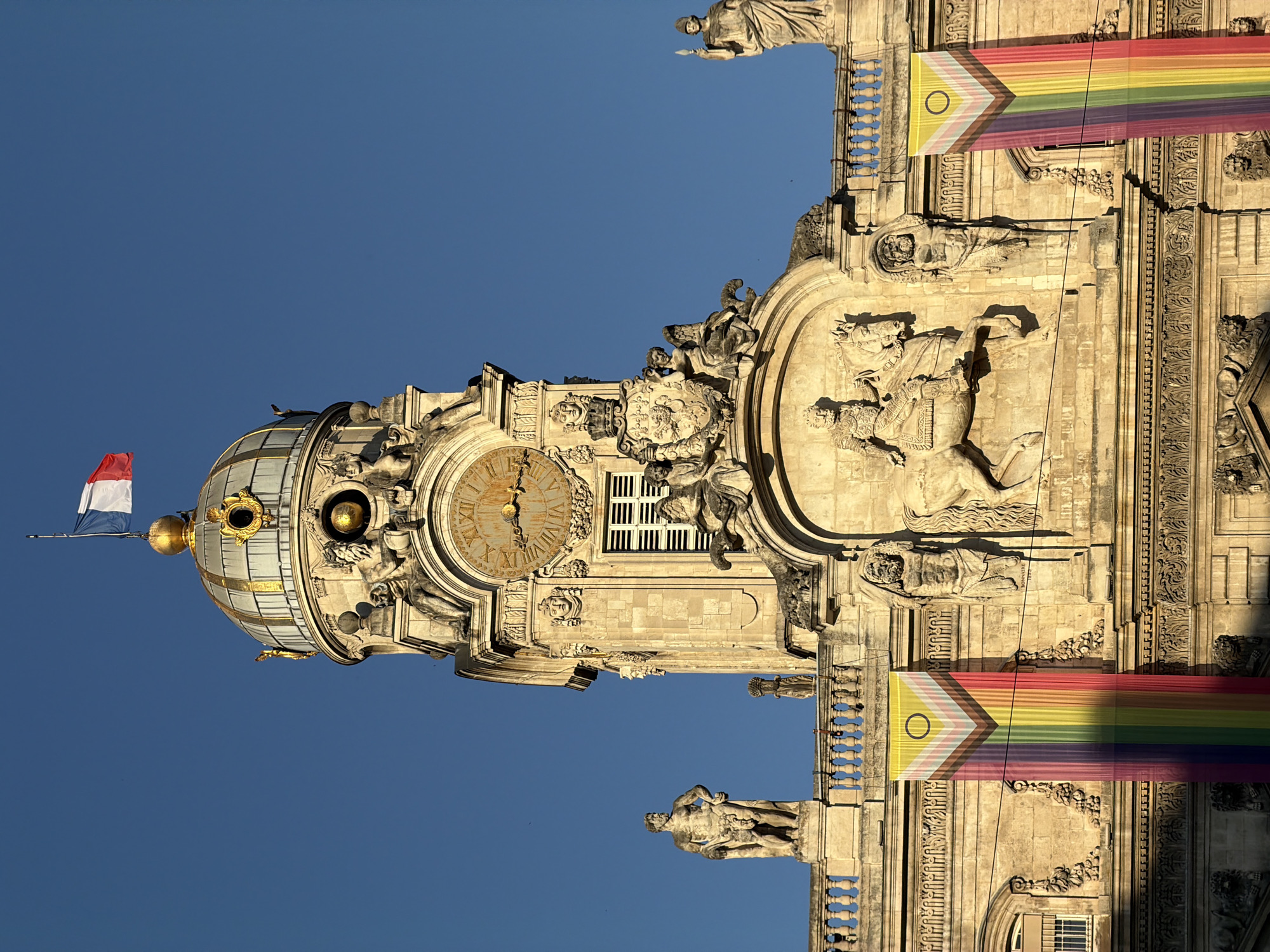 The image shows a close-up, upward-facing view of the Hôtel de Ville (City Hall) in Lyon, France, under a clear blue sky. The ornate stone building features a prominent clock tower with a golden dome topped by a French flag. The clock face has Roman numerals. Sculptural details, including statues and reliefs, adorn the facade. Two large banners, displaying a rainbow motif with a chevron pattern and a circle symbol, hang from the building's upper level, on either side of a central relief depicting a man on horseback. The architecture is classical and grand, indicative of a significant civic building. The sun is shining, casting shadows and highlighting the architectural features.
