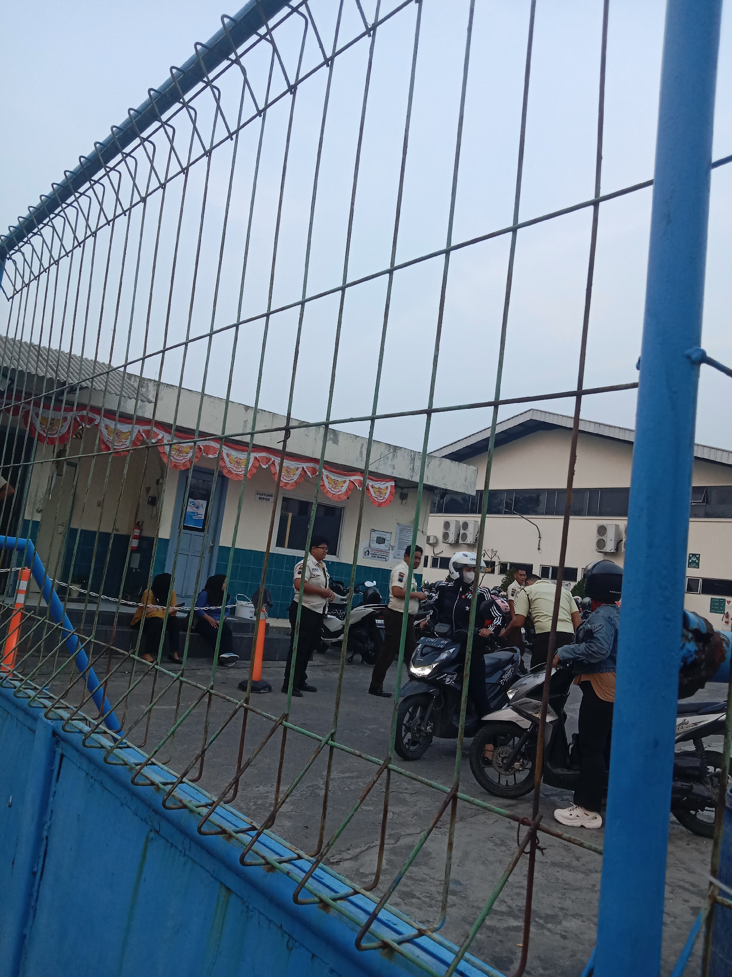 The image captures an outdoor scene at what appears to be a customs office or checkpoint. In the foreground, a blue metal fence with wire mesh obstructs the view. Beyond the fence, several people are gathered, some on motorcycles. A building with a white facade and a dark roof is visible, decorated with red, white, and orange banners. The building has windows and air conditioning units mounted on its exterior. Orange traffic cones are placed on the ground. People in various attire, including some in what appear to be uniforms, are interacting with each other or waiting near motorcycles. The overall environment suggests a functional area, possibly a border crossing or a transit point, under an overcast sky. The presence of a "CUSTOMS OFFICE" sign further confirms the location's purpose. The image does not provide specific details to confirm it is located in Semarang, Indonesia, but the visual cues are consistent with a port or border facility.