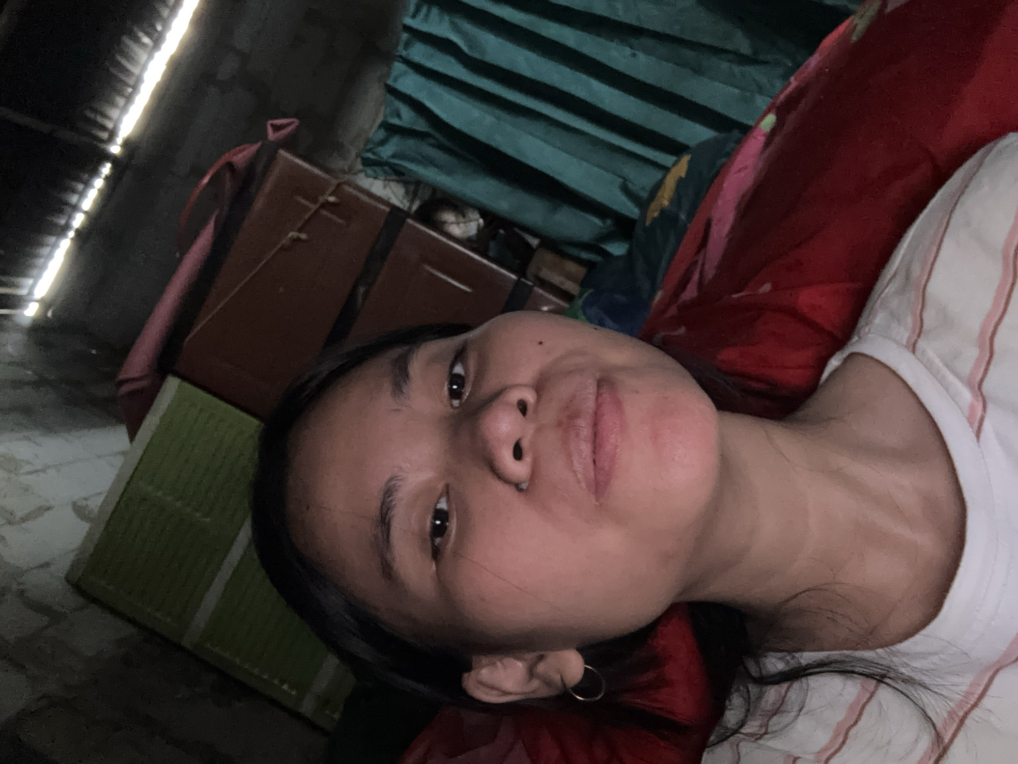 A selfie taken from a low angle shows a young woman lying down with her head propped up. She has dark hair and is wearing a white t-shirt with pink stripes and a hoop earring in her left ear. Her expression is neutral, with her mouth slightly open.
The background appears to be an indoor space with a rough ceiling and walls, possibly a storage area or makeshift dwelling. In the upper left, a green rectangular object is visible, possibly a mattress or a folded piece of fabric. To the right of that, a dark reddish-brown piece of furniture with latches is present. Behind the woman, a dark teal curtain is drawn, with some fabric peeking out from behind it. Sunlight streams in from gaps in the ceiling, creating bright lines. The overall impression is of a dimly lit, somewhat cluttered space.
The image's metadata suggests it was taken in East Jakarta, Indonesia, on May 19, 2024, at 10:19 AM.
