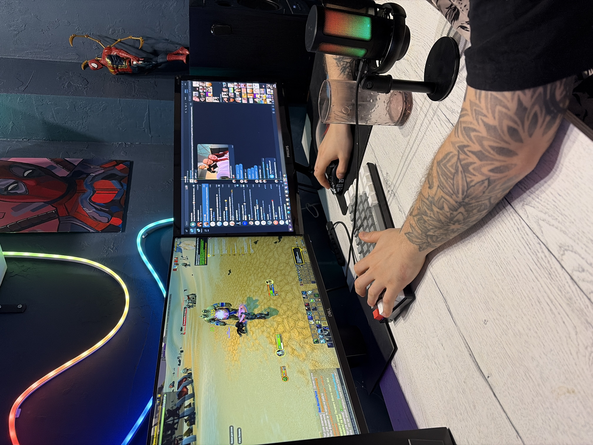 Here is a detailed description of the image:
**Scene:** The image shows a setup typically used for gaming or online communication, likely in a home environment.
**Objects:**
* Two monitors are stacked vertically. The top monitor displays what appears to be a chat application or streaming platform with various messages and avatars. The bottom monitor shows gameplay from the game World of Warcraft.
* A microphone with rainbow-colored lighting effects is positioned near a drinking glass.
* A keyboard and mouse are present, along with a mousepad.
* On the wall, there's a Spider-Man action figure and a decorative artwork that could be related to Deadpool. RGB LED light strips are also visible, adding ambient lighting.
**People:**
* A person's arm is visible with extensive tattoos, suggesting the person is seated at the desk using the gaming setup.
**Actions and Interactions:**
* The person's hands are positioned to use the mouse and keyboard, indicating active engagement with the computer setup.
**Environment:**
* The desk has a light wood grain finish.
* The wall is painted in a textured dark color, likely dark blue or gray.
**OCR Extract:**
* The OCR data from the screen mentions terms like "Чат для новичков" (Chat for Newbies), "Вэлтар-Ревуши ифибра," and dialogue related to the World of Warcraft game, confirming the active use of the gaming setup. The OCR also contains text in English, indicating the multi-lingual use of the computer.
The image does not provide enough information to indicate the city of Rostov-on-Don, Russia.