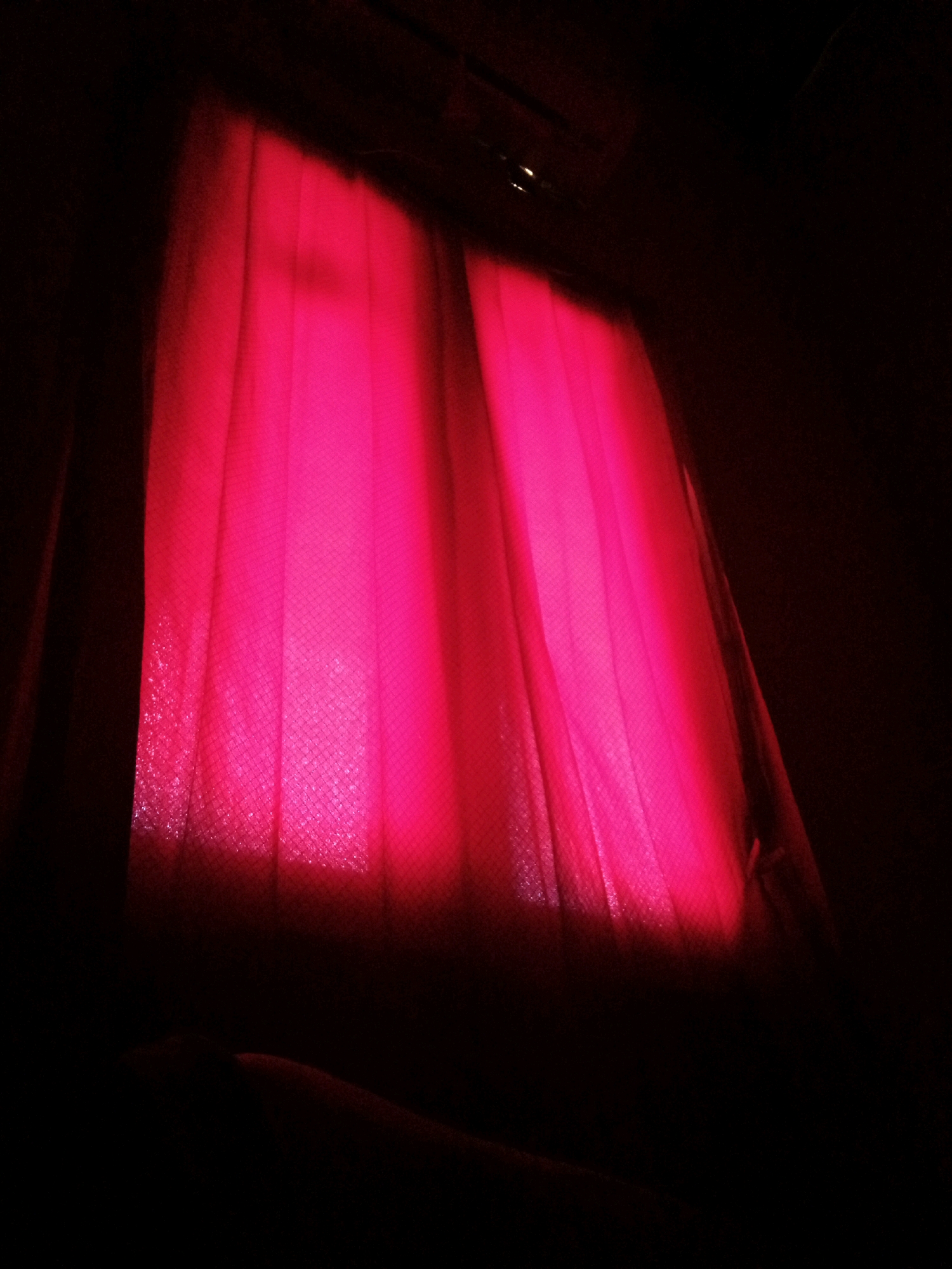 The image shows a close-up of vibrant pink curtains illuminated by an unseen light source. The curtains are gathered, creating vertical folds, and appear to be made of a textured fabric, possibly with a subtle diamond pattern. The lighting is intense and diffused, casting a strong pink glow on the fabric and obscuring any details of the environment beyond. The top and right sides of the image are dark, suggesting the curtains are indoors and potentially covering a window or a stage. There are no people or animals visible in the scene. The image does not provide any information about the city of Pelaihari, Indonesia.