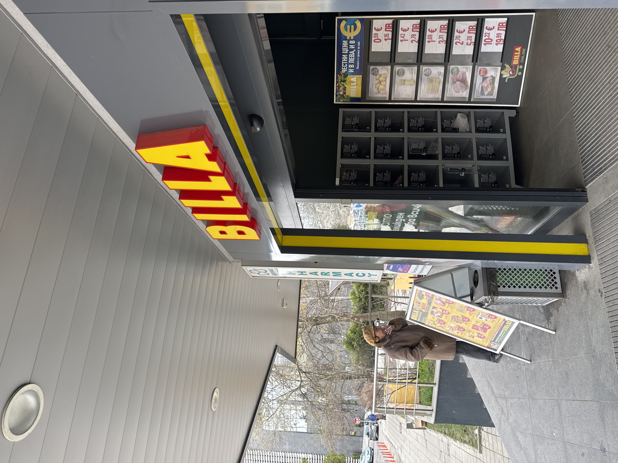 The image shows the exterior of a Billa supermarket, a pharmacy sign (in Bulgarian and English) and a woman standing on the sidewalk in Sofia, Bulgaria. The Billa signage is prominent, with the store's name in large yellow and red letters above the entrance. A digital screen, with various prices and options (in Bulgarian and English) for "Billa Online" food purchases, is beside the door. A sandwich board displays advertisements. The woman is wearing a coat, a head covering and stands beside a trash bin and a small promotional display for Billa. The surrounding environment includes trees, a building, a street with parked cars, and a glimpse of other buildings.