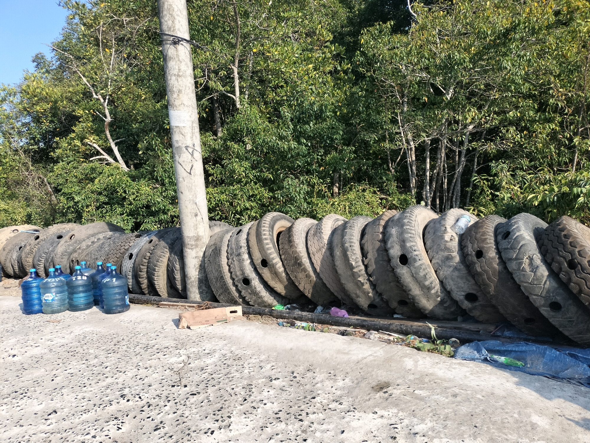 In the city of Bunati, Indonesia, a scene of discarded tires and water jugs is captured. The image, taken outdoors during daylight, showcases a row of large, used tires stacked against a backdrop of lush green trees. A concrete pole stands prominently to the left, with black wires attached. In the foreground, several blue water jugs are arranged on a concrete surface, some of them appearing to be full. To the right of the jugs, a cardboard box is visible, and beyond that, more tires are stacked in a semi-circular formation. The ground is a textured concrete, with scattered debris and a blue tarp visible further down. The overall impression is one of an informal storage or disposal area for waste materials in a semi-rural or industrial setting.