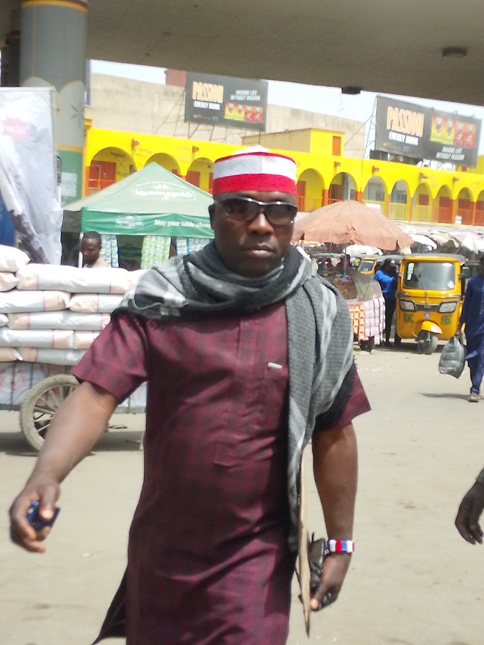 The image depicts a daytime street or market scene in Fagge C, Nigeria. In the immediate foreground, an adult male, appearing middle-aged, is walking forward, facing the camera. He wears a red and white striped traditional cap, dark sunglasses, a grey and black patterned scarf, and a short-sleeved maroon tunic with a breast pocket. His right hand holds a small dark blue object. His left hand holds a flat, light brown rectangular object and a dark wallet/bag, and he wears a red, white, and blue striped wristband.
In the midground to the left, a wooden two-wheeled cart is laden with numerous white sacks. Behind this, a green tent with partial white lettering, possibly reading "May your table always," is visible. A person is partially obscured behind the tent.
In the midground to the right, a yellow three-wheeled vehicle (auto-rickshaw) is parked. An orange parasol provides shade over an indistinct stall. Another individual in a blue garment is seen walking away, carrying a black bag.
The background features a long, yellow building with multiple arched entrances and red-orange doors, suggesting a market or commercial structure. Above this building, two large billboards display "PASSION ENERGY DRINK" with the slogan "MAKE LIFE SWEET WITHOUT PASSION." The ground is an unpaved or dusty surface.