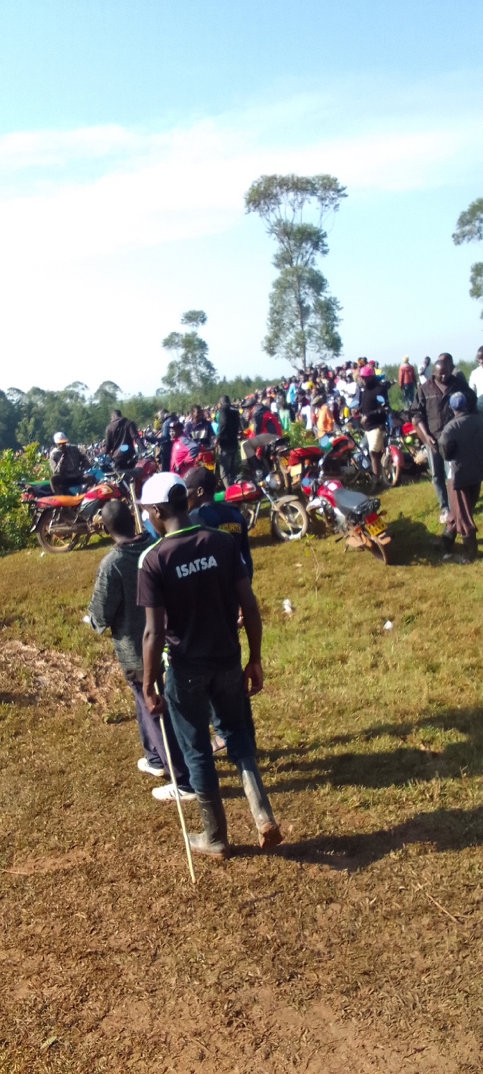 The scene is a crowded gathering of people near a dirt road in Isukha ICHINA, Kenya. A large group of people are standing along the dirt road and on the grassy slope. A number of motorbikes are parked in a line along the road. Two men wearing black, blue, and white clothes are standing in the foreground. The man on the right is wearing a black shirt with white lettering that reads "ISATSA". Both men are looking toward the group of people. The ground is covered in brown grass and dirt. The sky is clear with a few white clouds. There are trees in the background. The area is sparsely populated. The scene is peaceful and orderly.