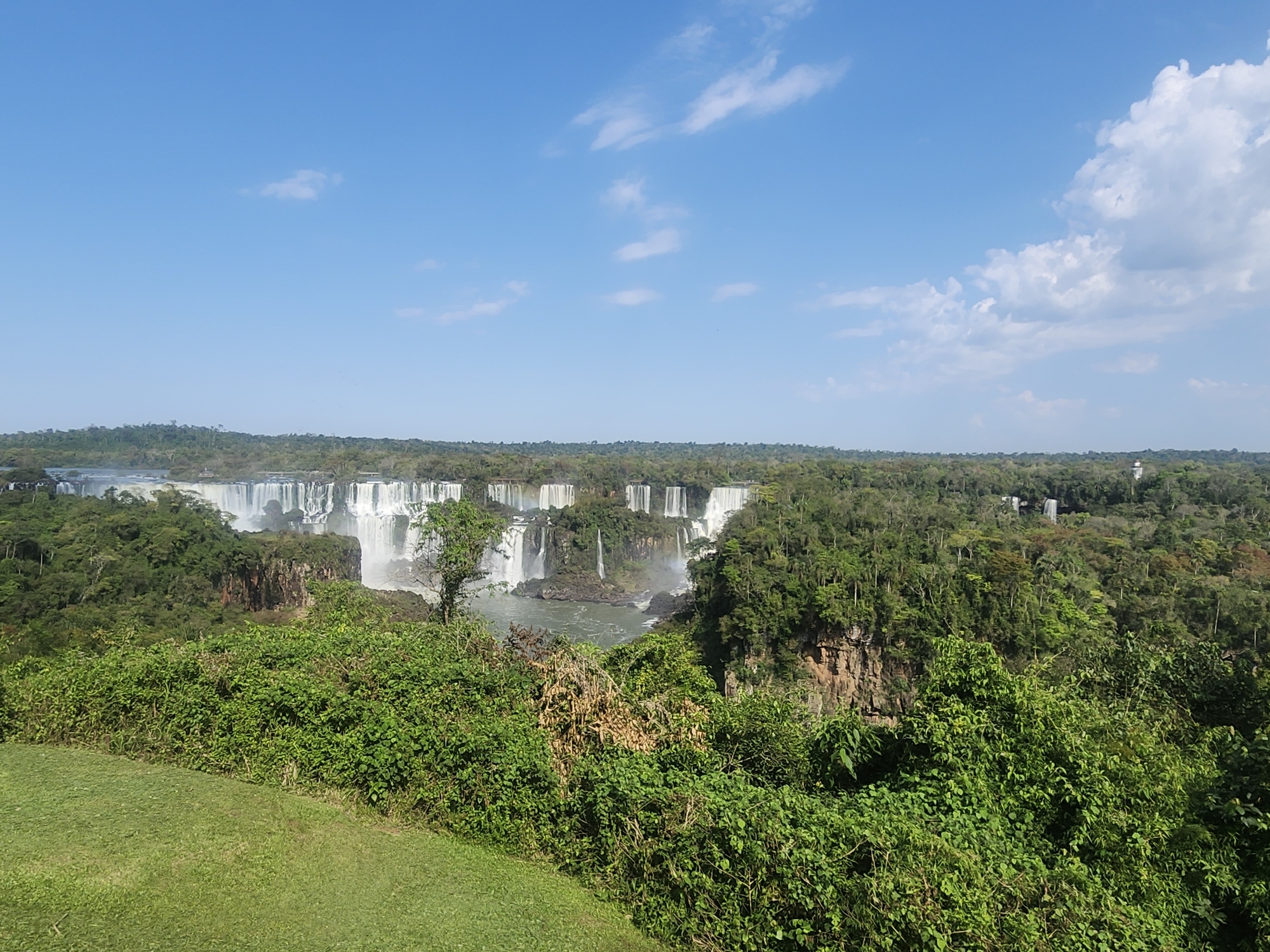 The image depicts a panoramic view of Iguazu Falls, located in Iguazu National Park, Foz do Iguaçu, Brazil. A vast expanse of lush green forest frames the majestic waterfalls, which cascade over a wide, rocky cliff face. The water plunges into a turbulent river below, creating a mist that rises into the air. The sky is a clear blue with scattered white clouds. In the foreground, a neatly mown green lawn slopes gently upwards, bordered by dense green foliage and shrubbery. The scene is serene and awe-inspiring, showcasing the raw beauty of nature.