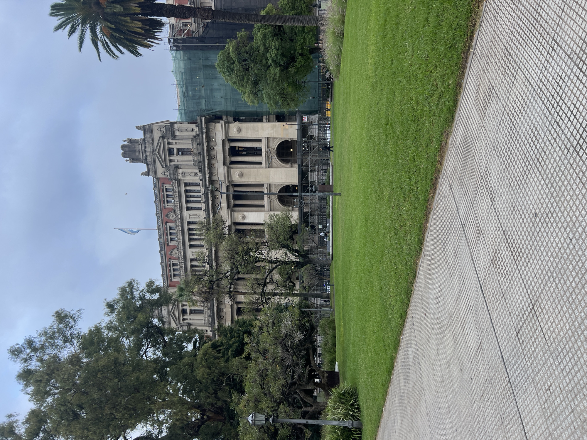 The image depicts a street-level view of the Palace of Justice in Buenos Aires, Argentina. The building is visible behind trees and a lawn, and is partly covered with scaffolding. A flag of Argentina is waving atop the palace. A paved pathway is in the foreground of the image.