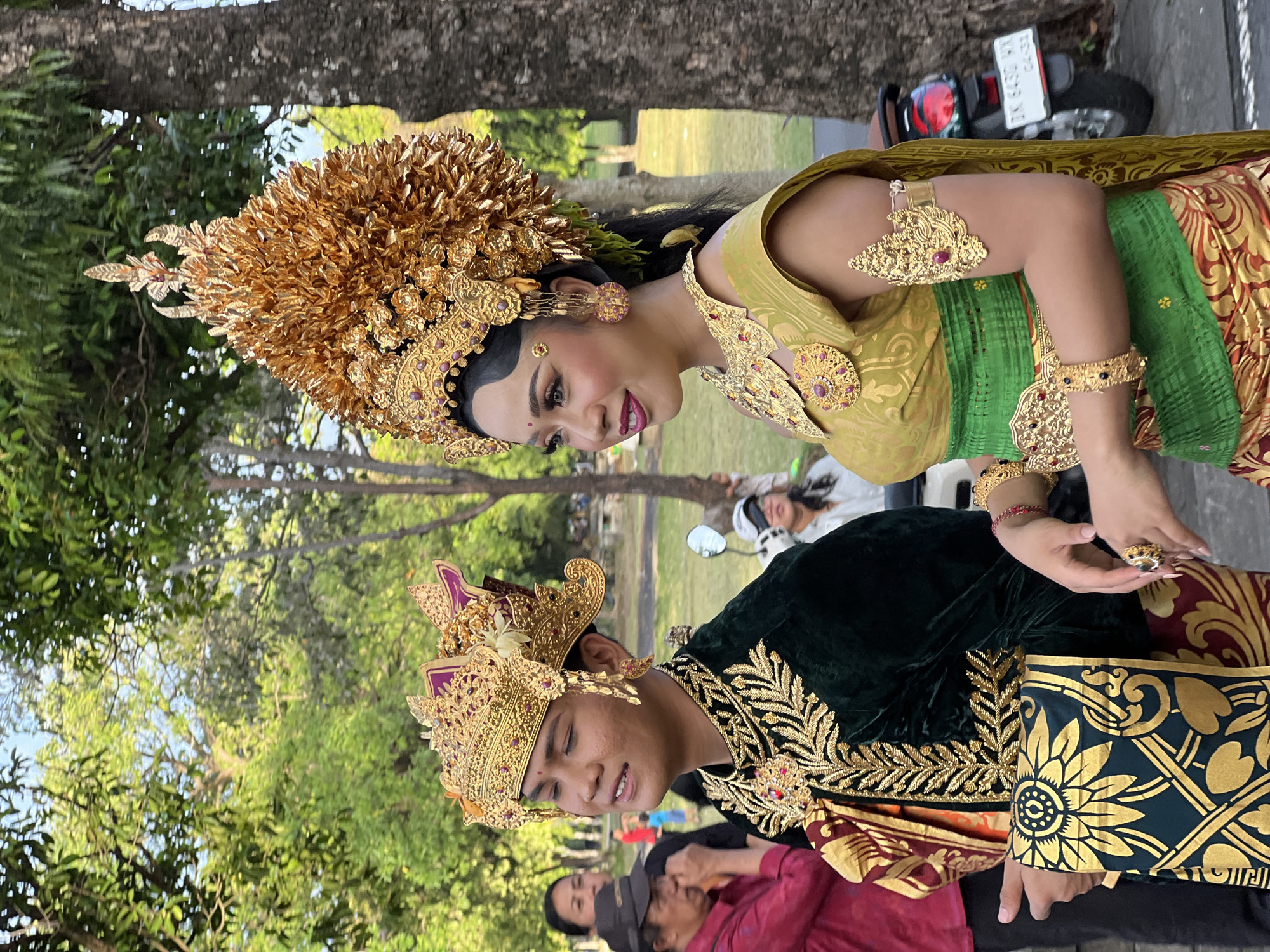 Two individuals, a male and a female, are posing for a photograph outdoors, likely in a park or public space within Denpasar, Indonesia. Both are adorned in elaborate traditional Balinese attire, characterized by rich gold ornamentation, vibrant fabrics, and ornate headdresses.
The male subject, on the left, wears a dark green velvet jacket with intricate gold embroidery around the collar and down the front. He has a matching gold headdress with feather-like protrusions and a prominent red jewel at its center. A patterned cloth, dark green with gold floral motifs, is draped over his arm. He is looking to his left with his eyes closed and his mouth slightly open.
The female subject, on the right, is wearing a gold and green outfit. Her headdress is a large, golden, tiered structure, also featuring intricate designs and what appear to be small green accents. She is adorned with numerous gold jewelry pieces, including a large necklace, bracelets on both arms, and earrings. Her outfit includes a green sash tied around her waist. She is looking to her left with a slight smile.
In the background, other people are visible, some blurred, suggesting a public gathering or event. Trees and greenery form the backdrop, indicating an outdoor setting. A parked motorcycle with a license plate is visible to the right of the female subject. The overall scene suggests a cultural event or performance taking place in Denpasar.