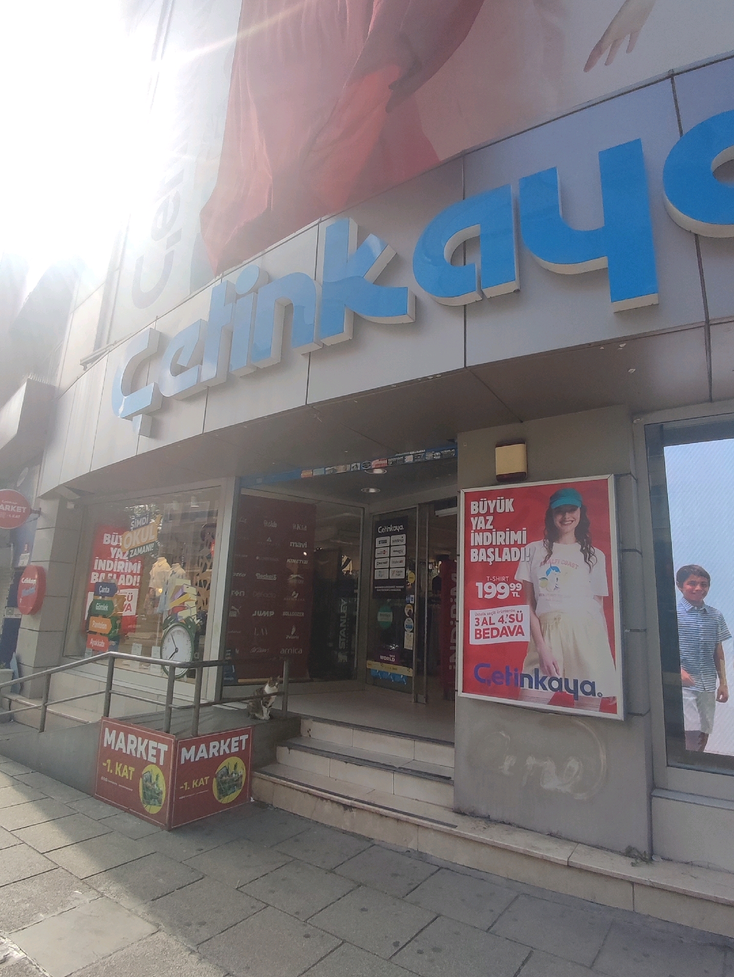 The image shows the exterior of a "Çetinkaya" store in Kadıköy, Turkey. The store's name is prominently displayed in large blue letters across the facade. In the foreground, there are two red signs that say "MARKET -1. KAT," indicating a market on the first basement level. A cat is perched on a railing to the right of the entrance.
Through the store windows, various branded items and promotional materials are visible. A large advertisement featuring a young woman wearing a t-shirt and cap is displayed on the right side of the building, announcing a "BÜYÜK YAZ İNDİRİMİ BAŞLADI!" (Big Summer Sale Has Started!). Another advertisement on the same poster states "T-SHIRT 199,99 TL" and "3 AL 4.SÜ BEDAVA" (Buy 3, Get the 4th Free). To the right of this advertisement, a young boy in a striped shirt and shorts is visible looking towards the camera through a display window.
The building appears to be a commercial establishment on a street with a sidewalk made of grey paving stones. The lighting suggests it is daytime, with the sun casting a bright glare on the upper part of the facade.