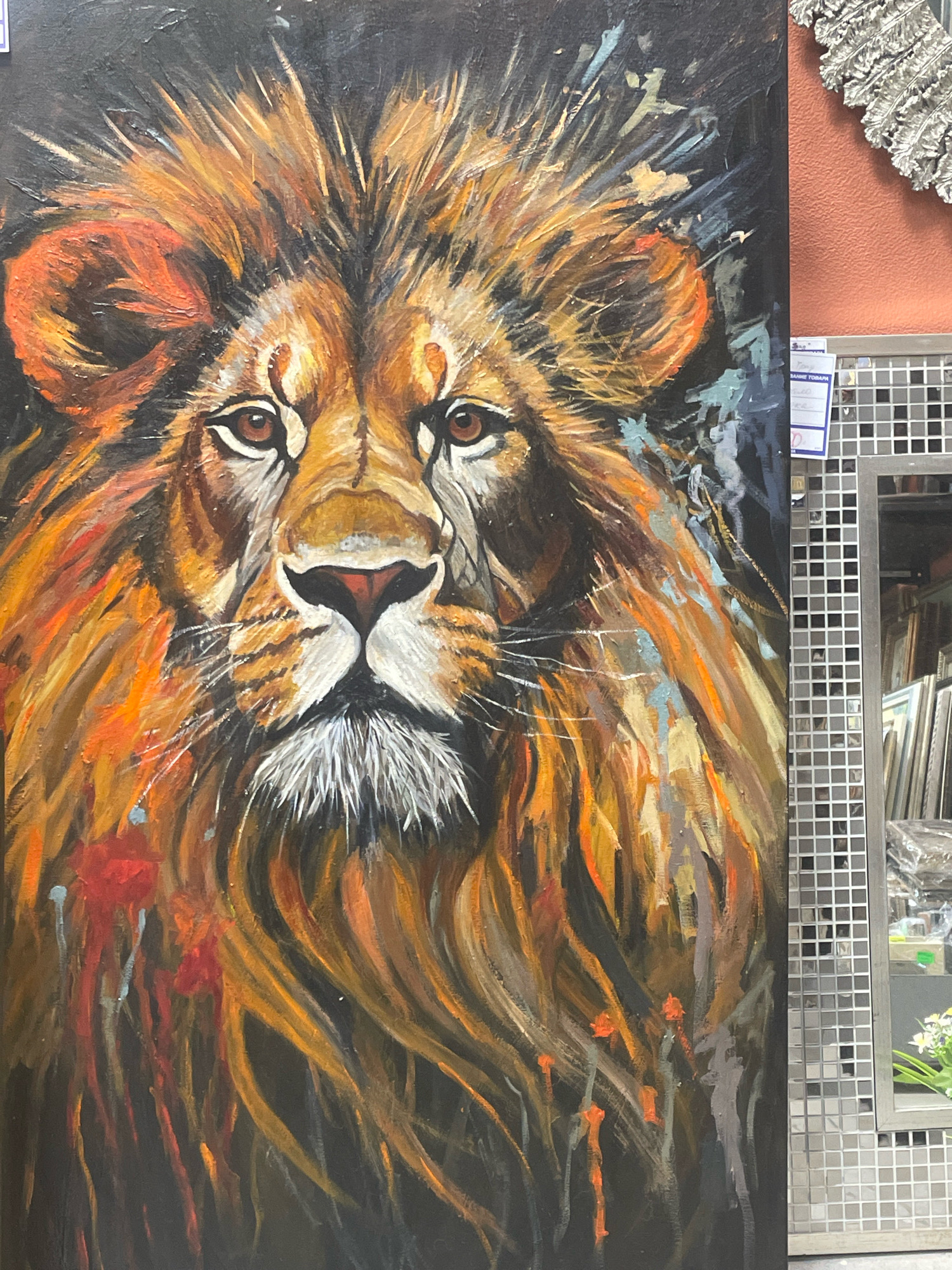 A close-up, vertical, eye-level shot depicts a vibrant, textured oil painting of a lion's face. The lion's mane is rendered in bold strokes of orange, yellow, and black, creating a sense of wild energy. The lion's eyes are brown and intense, and its nose is a striking red and black. Splatters of red and orange paint are visible on the left side of the canvas, adding to the dynamic feel of the artwork. The background is a dark, textured black.
To the right of the painting, a silver, mirrored mosaic tiled column rises vertically. Behind the column, a muted coral-colored wall is visible. Further to the right, a glass-fronted display case with multiple shelves is present, showcasing various framed items, likely artwork or photographs, on display. The floor appears to be tiled with large, grey squares.
The artwork is displayed in what appears to be a commercial establishment, possibly an art gallery or frame shop, judging by the display of other framed items. The context of Saint Petersburg, Russia, is inferred from the presence of Cyrillic text on a white tag attached to the display case, which reads in part "ВАЖНЕЙШИЕ ТОВАРЫ" (Vazhneyshiye tovary), meaning "Most Important Goods." The overall impression is that of an art retail environment.