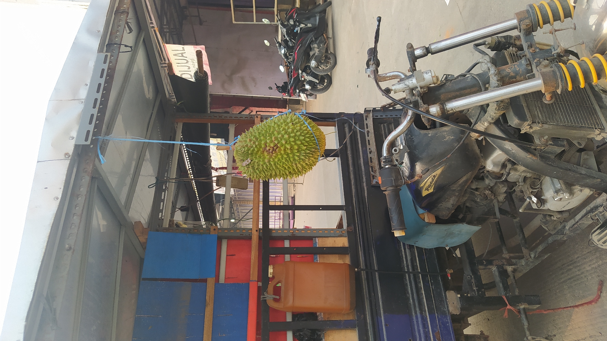 This image depicts a street scene in Klapanunggal, Indonesia, featuring a three-wheeled vehicle that appears to be a modified motorcycle with a cargo bed. A large, spiky durian fruit is suspended by a blue cord from the vehicle's roof structure, which is made of corrugated metal and metal framing. To the left of the durian, a brown plastic jerrycan is also hanging from a hook. The cargo bed of the vehicle contains stacked red and blue boxes, suggesting it is used for transport or as a mobile vendor. In the background, several motorcycles are parked, and a sign with the word "DIJUAL" (for sale) is visible. The ground is paved with concrete. The overall environment suggests a commercial or marketplace area within the city.