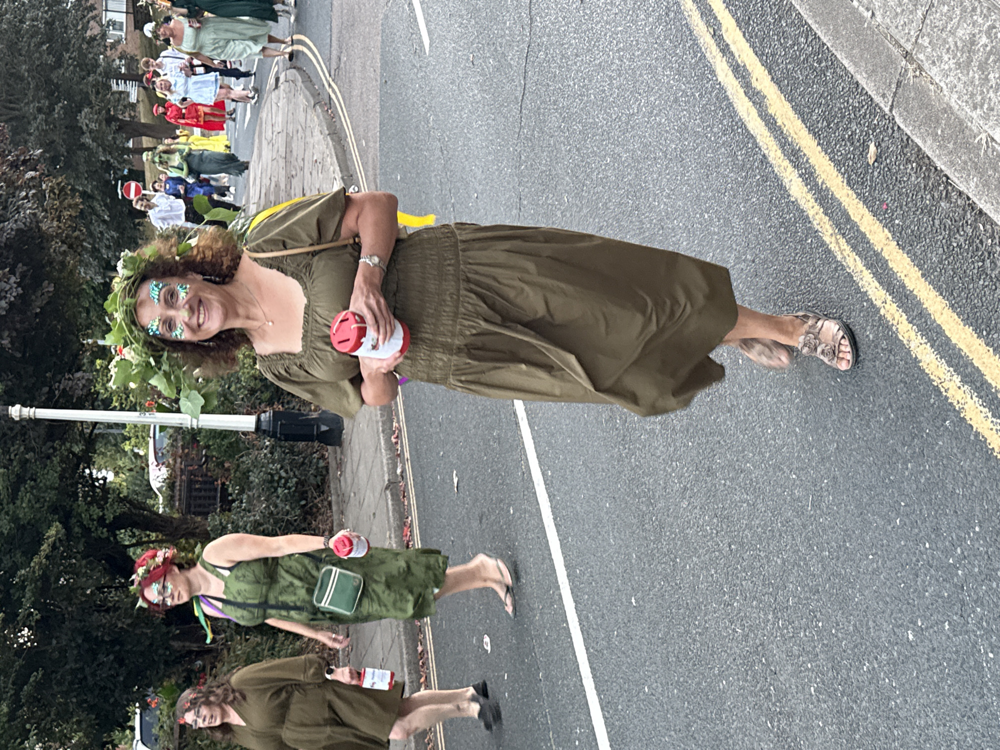 In St Leonards, United Kingdom, a group of people are participating in a parade or festival. The foreground features a woman wearing a green, smocked dress and a floral wreath on her head, adorned with glitter. She is holding a red and white collection tin. To her left, another woman in a similar green dress with a red floral headband and glitter on her face is also walking and holding a collection tin. Further back, other participants are visible, some in green attire, and others in colorful outfits including red and yellow. The scene takes place on an asphalt road with yellow double lines, bordered by a sidewalk and some greenery. The overall atmosphere suggests a celebratory event.