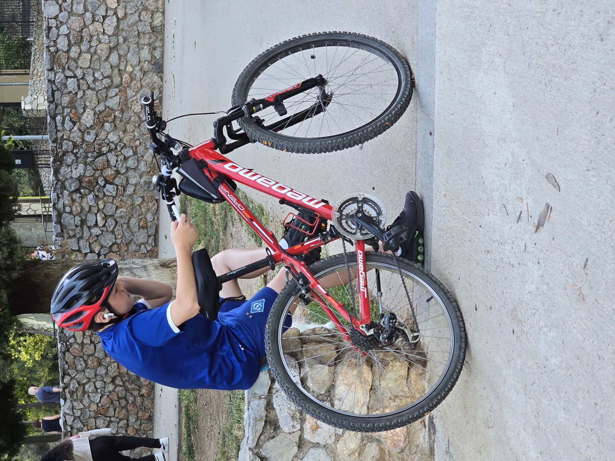 A young person wearing a blue shirt, black shorts, a red and black helmet, and white earbuds is sitting on a stone wall next to a red bicycle. The person is looking down at their hands, which are holding a mobile phone. The bicycle is a mountain bike with knobby tires and a black water bottle cage. In the background, there is a stone wall, trees, and other people walking. The scene appears to be in a park. The location in the city of Cornellà De Llobregat, Spain, is a public park with a paved walkway and a natural stone wall.
