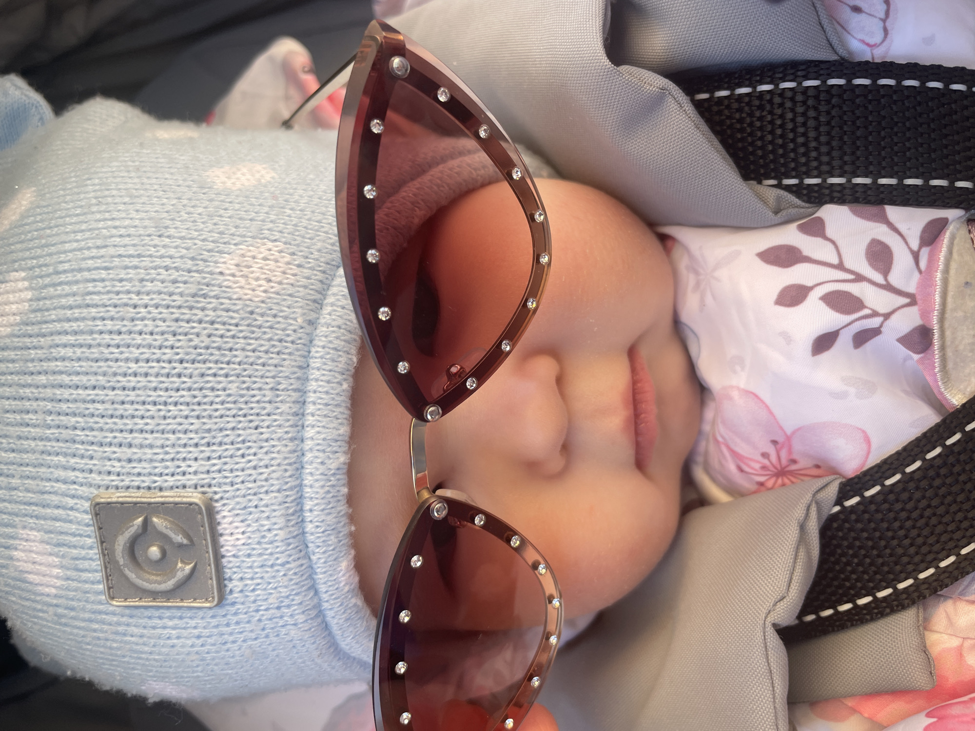 A baby wearing a blue knit hat with a logo lies in a carrier. A pair of rose-colored sunglasses is placed on the baby's face. The carrier has gray fabric and a floral-patterned lining. A black strap is also visible. The image appears to be taken indoors. There is no evidence indicating the location is in the city of Chaikovsky, Russia.
