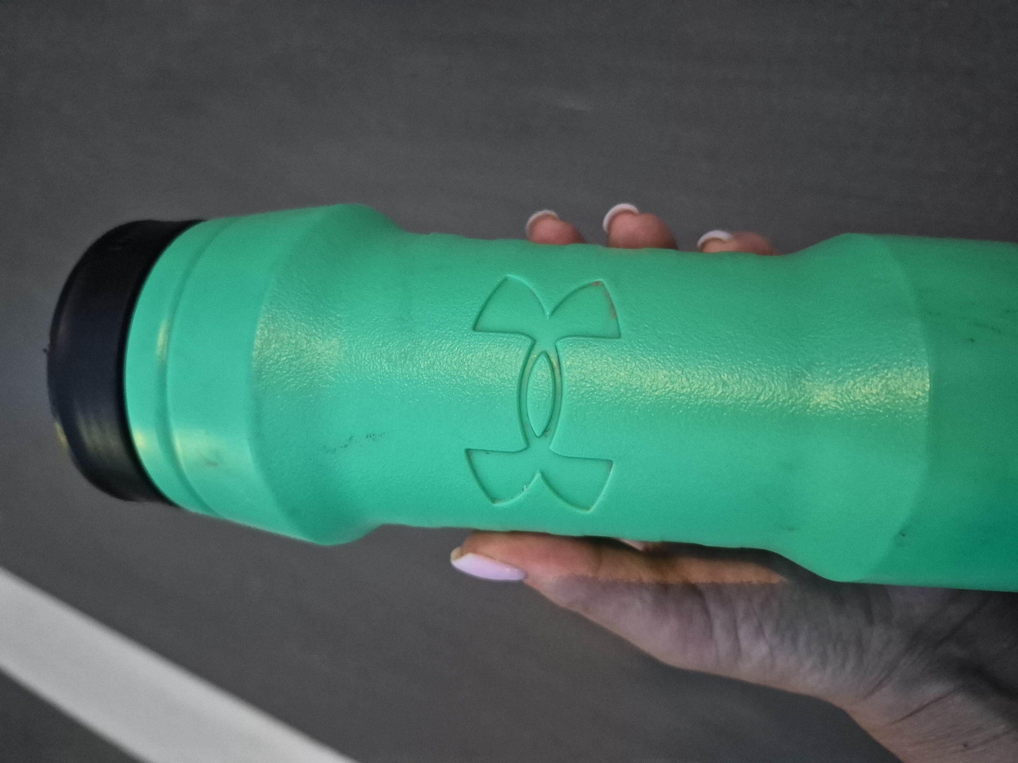 A close-up, angled shot shows a bright green water bottle, possibly plastic, with the Under Armour logo embossed on its side. The logo is a stylized "UA" symbol in a darker shade of green, suggesting it's part of the bottle's design rather than a separate sticker. The bottle has a black cap and is held by a person's hand, which is partially visible in the lower right of the frame. The hand has light skin with pink nail polish on the visible fingernails. The background is a muted gray, with a white stripe running diagonally from the bottom left corner towards the right. The lighting appears to be natural, possibly from an overhead source, given the way it highlights the curve of the bottle and casts subtle shadows. The focus is sharpest on the bottle and the logo, with the background slightly blurred, indicating a shallow depth of field. No text is visible on the bottle or in the background. The image does not provide specific clues about the time of day, weather, or location, other than suggesting an indoor or shaded outdoor environment. The overall impression is that of a simple product shot, highlighting the water bottle and its branding.