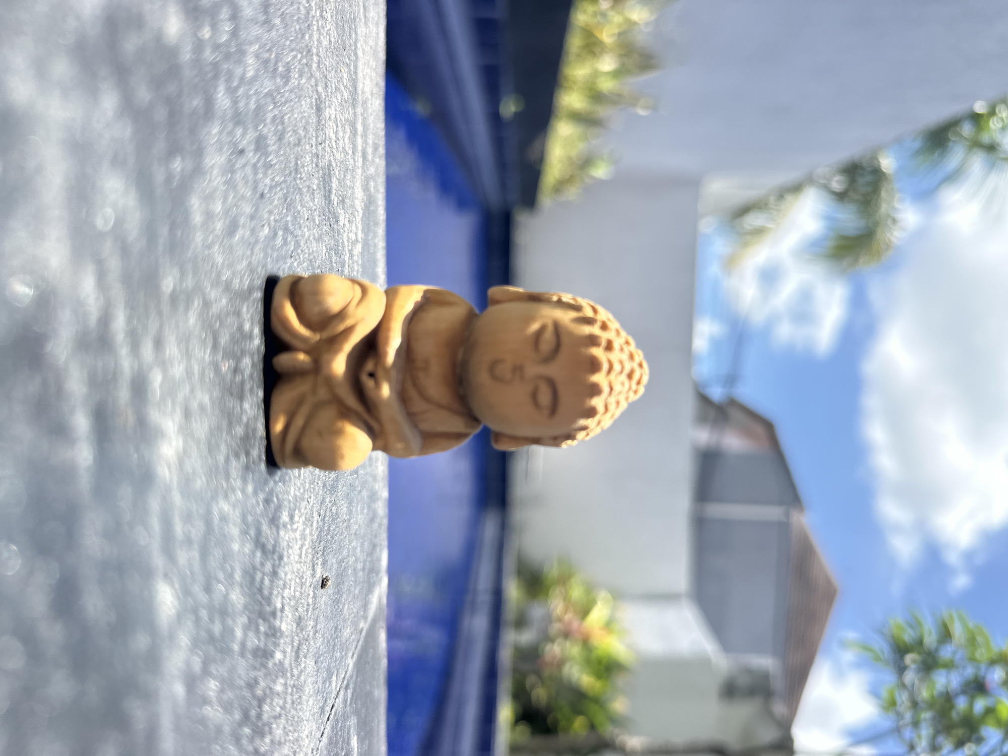 A small, light brown wooden Buddha statue is the central subject, positioned horizontally on a textured gray surface. Behind the statue, a blue structure is visible, possibly a part of a pool or similar feature. In the background, blurred elements suggest foliage, a building with a gray roof, and a blue sky with clouds. The location is likely Dalung, Indonesia, based on the user's prompt. No people, interactions, or specific actions are discernible in the image.