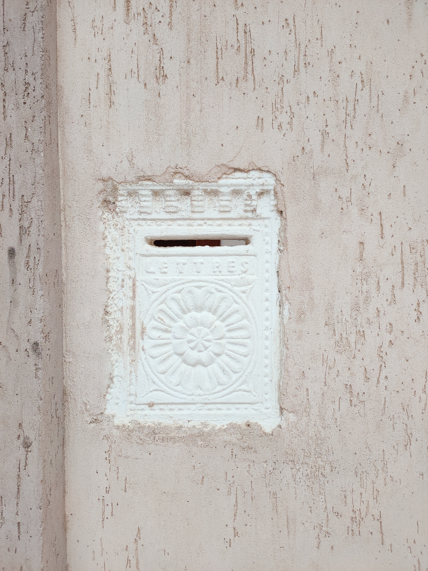 This image shows a close-up of a white, ornate letterbox embedded in a textured, beige wall. The letterbox is rectangular with decorative elements, including a slot for mail at the top and a circular, floral-like relief below. The word "LETTRES" is visible in raised lettering above the floral design. The wall surrounding the letterbox has a rough, stucco-like finish with visible vertical streaks and some signs of wear and tear, particularly around the edges of the letterbox. The overall impression is of an older, perhaps historic, architectural feature. The location in the city of Ariana, Tunisia, is not explicitly visible in the image itself but is provided as context.