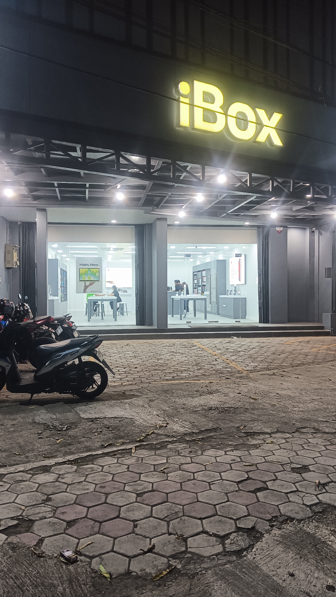 The image depicts the exterior of an iBox store, a retail outlet for Apple products, located in Sragen, Indonesia. The store's prominent yellow neon sign with the "iBox" logo is clearly visible above the entrance. The storefront features large glass windows and doors, revealing the interior which is illuminated and displays various Apple products and displays. Inside, two individuals can be seen interacting with displays or staff. In front of the store, several parked motorcycles are present on a cobblestone pavement. The lighting suggests the scene is captured at night.