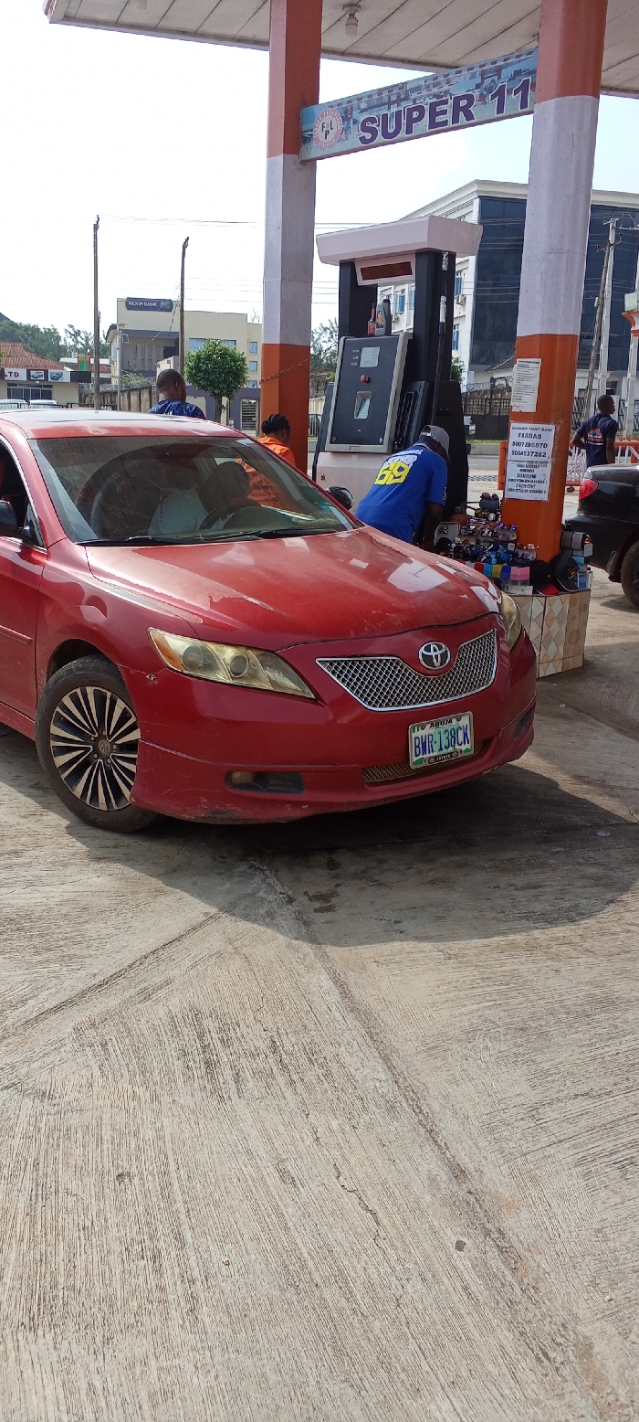 The image depicts an active scene at a gas station identified as "F & L SUPER 11" in Akure, Nigeria. A red Toyota Camry, license plate "ABUJA BWR-138CK", is parked on a rough concrete surface in the foreground, facing slightly left. A gas station attendant, wearing a blue shirt with "SUPER 69" on the back, is positioned at a black and white fuel dispenser, actively fueling the red vehicle. The car's driver is partially visible through the windshield. To the right of the fuel dispenser, a small makeshift retail display is set up on a tiled base, offering various items like sunglasses and hats. A pillar adjacent to this display bears a white sign listing "FARDAS" and multiple Nigerian bank account details. In the background, other individuals are present, including a person in an orange shirt near the fuel pump and another in a blue shirt further right, near a partially visible black vehicle. Beyond the gas station structure, a building marked "HEXIM BANK" is visible, along with other urban structures and sparse vegetation under a bright sky.