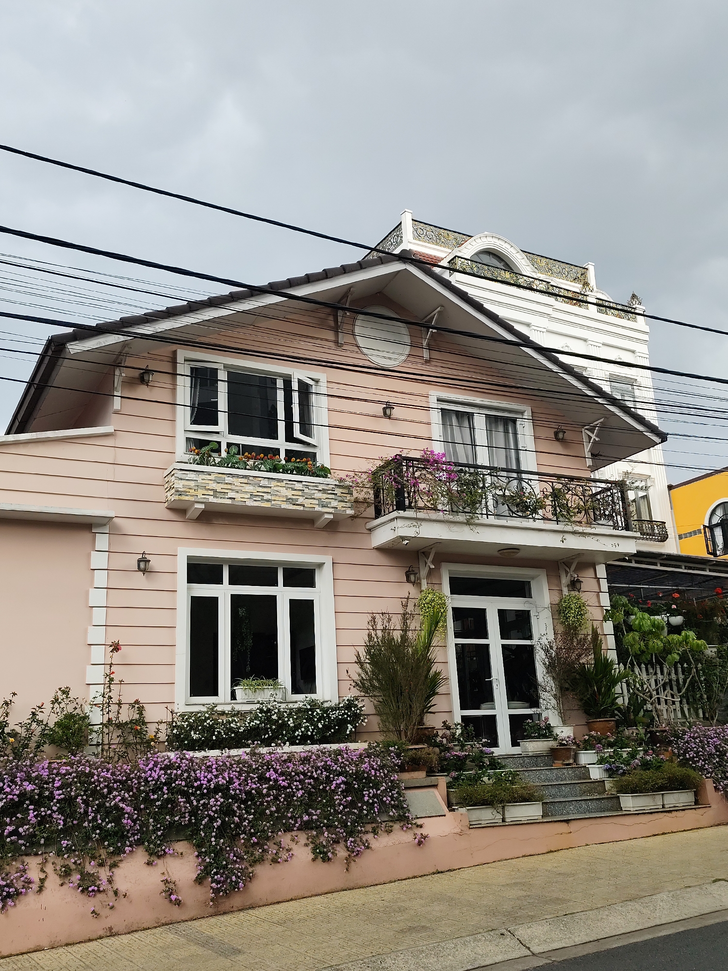 A two-story residential house with a pink façade is centered in the frame, exhibiting a Western architectural influence. The house features white-framed windows, a balcony with ornate black metal railings, and a gabled roof with a dark trim. Abundant greenery and flowers, including a dense cluster of purple flowers along the base of the house, enhance its appeal.
Behind the pink house, a taller, white building with decorative details is partially visible. Electrical wires crisscross the sky, and a glimpse of another yellow building is seen on the right.
The scene is set on a sidewalk with a street visible in the foreground, indicating an urban environment. The overall impression is of a well-maintained and aesthetically pleasing residential area. The image is in Da Lat, Vietnam.