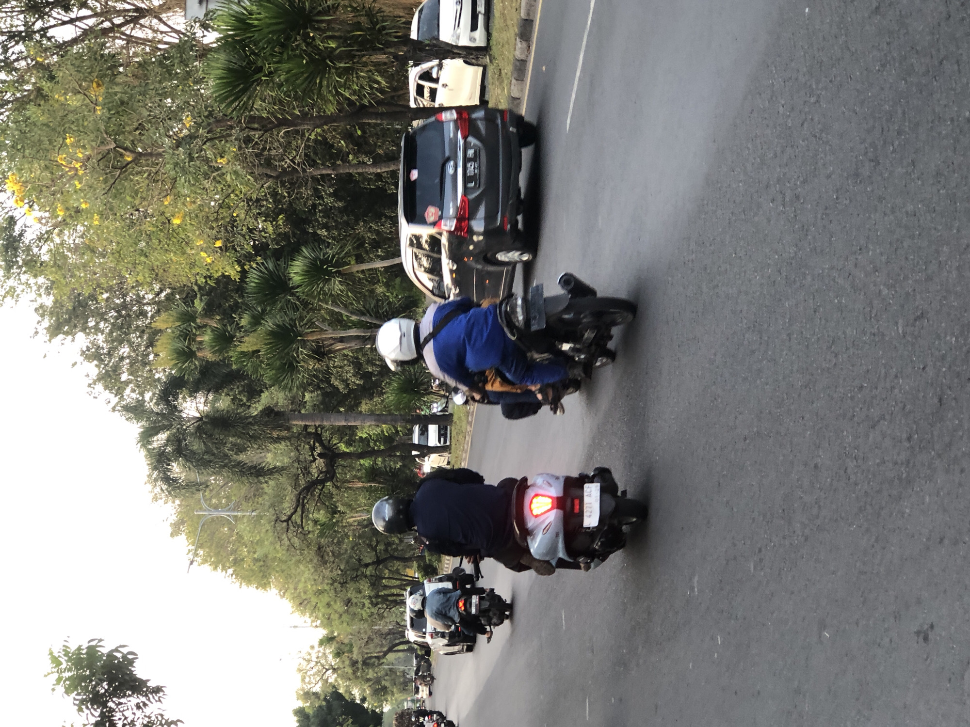The image depicts a street scene in Gelam, Indonesia, with multiple motorcycles and cars on a paved road. The setting appears to be during daylight, with trees lining the street and a bright sky in the background. Several individuals are riding motorcycles, some wearing helmets and casual clothing, while others are in what appears to be uniforms. One person is wearing a blue and white outfit, and another is in a dark uniform. Cars are also present, including a dark grey SUV with a license plate visible, and white cars further down the road. The road surface is asphalt, with lane markings visible. The overall impression is of a typical urban or suburban street with moderate traffic.