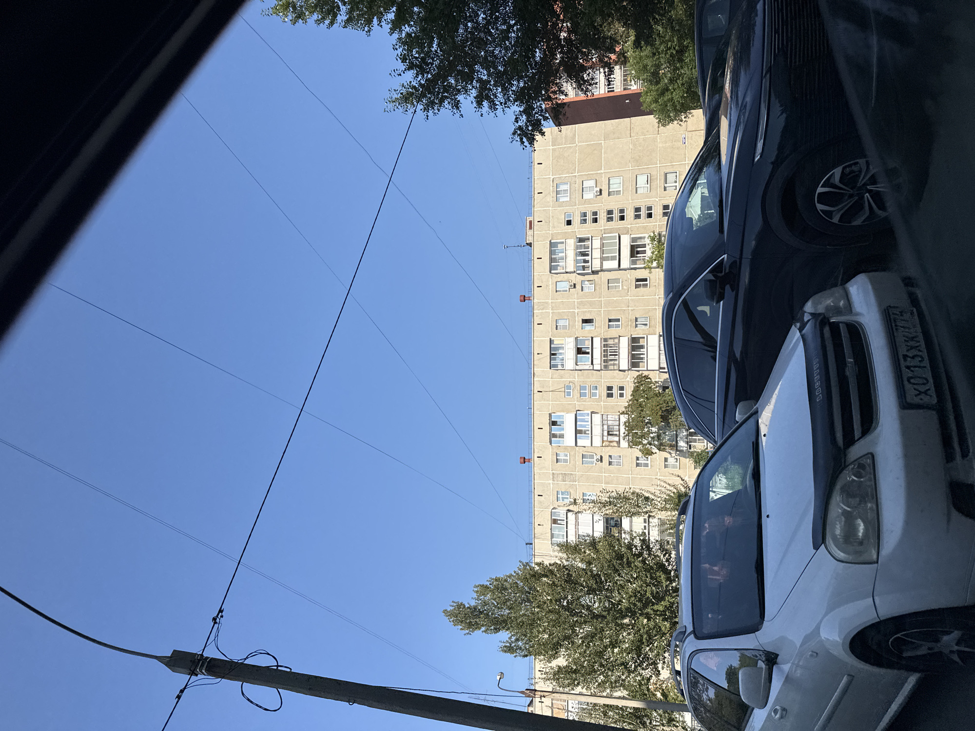 The image shows a street scene in Chelyabinsk, Russia, on a clear day. A tall, concrete utility pole with wires runs up the left side of the frame. In the foreground, two cars are parked: a white Chevrolet estate wagon on the left, and a dark-colored SUV on the right. Behind the cars is a multi-story, beige apartment building with many windows and balconies. Trees with green foliage are visible between the cars and the building, and also to the right of the building. The sky is a bright, clear blue, with several power lines crisscrossing the upper portion of the image.