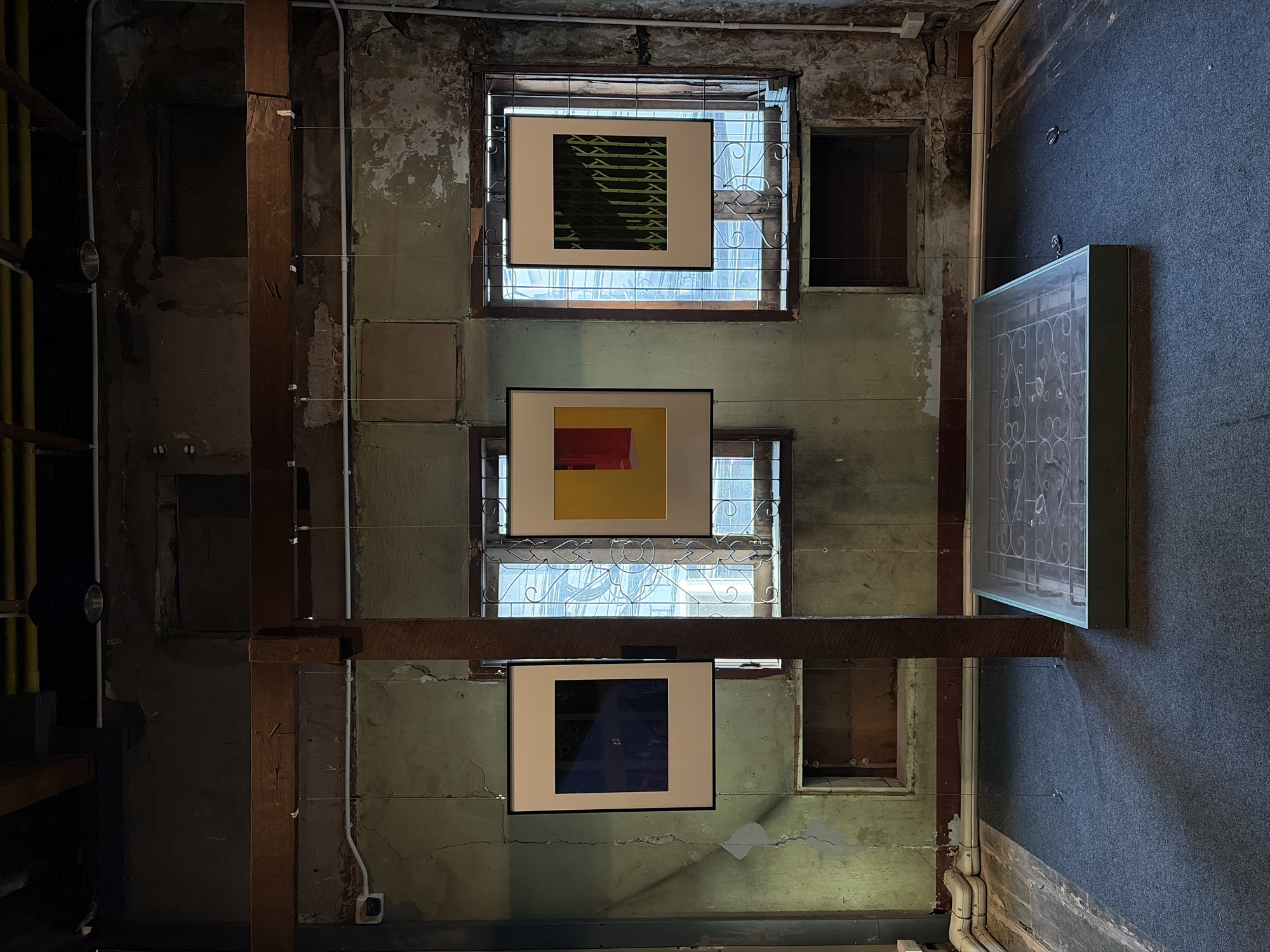 An interior shot of a building in Bangkok, Thailand. The walls are distressed, with peeling paint and exposed patches. Three framed artworks hang suspended in front of barred windows. The top artwork features green and black geometric patterns. The middle artwork is predominantly yellow with a red rectangle. The bottom artwork is mostly blue. A dark wooden beam runs horizontally across the middle of the frame, suspending the artworks. To the left, a dark, multi-tiered shelf structure is visible. To the right, a grey wall meets the distressed, light green wall, and a framed decorative panel hangs. White pipes run along the bottom right corner. The scene is dimly lit.