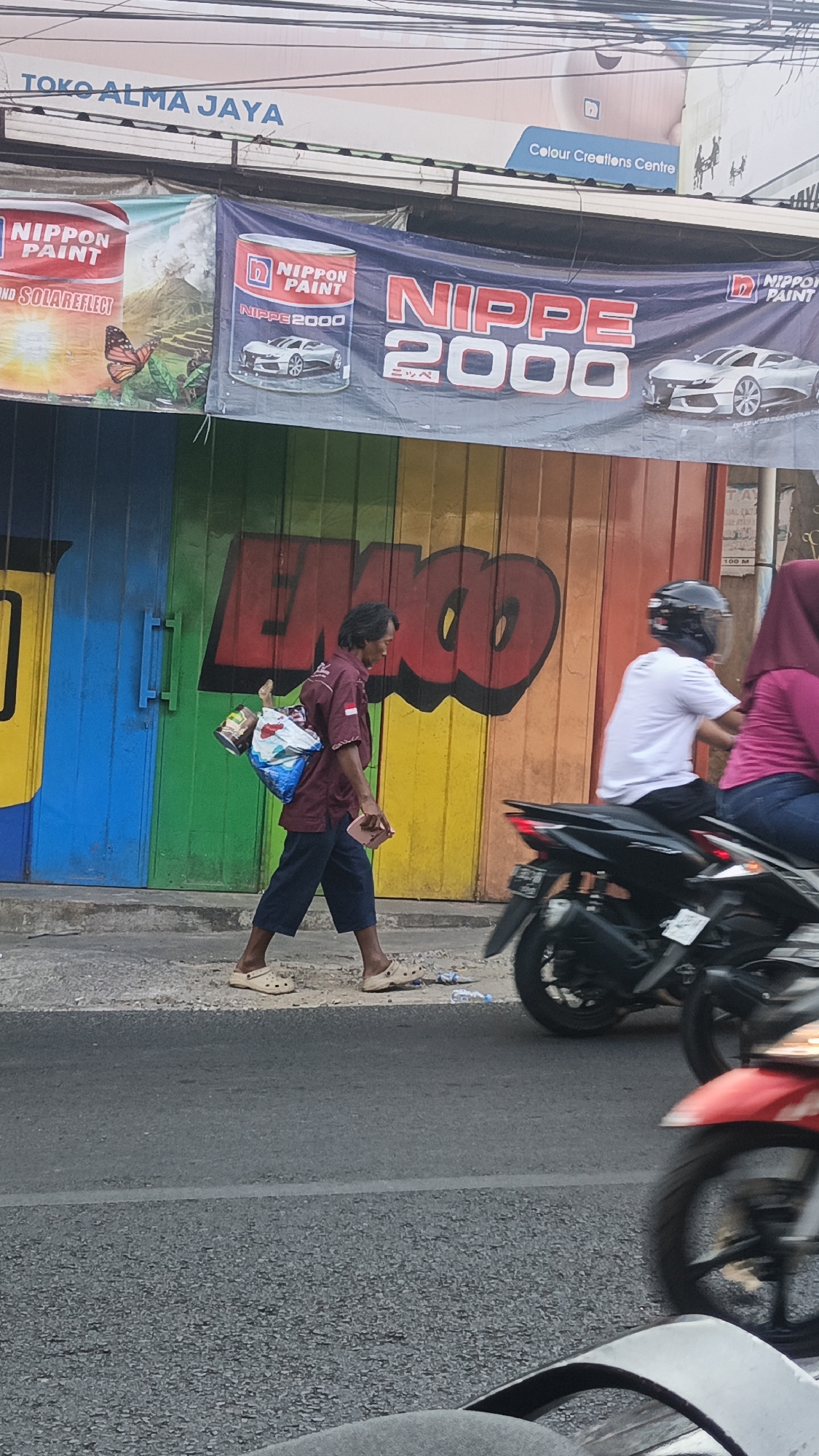A street scene in Dawe, Indonesia, shows a man walking on a sidewalk in front of a colorful, rainbow-striped garage door with graffiti that reads "EXOD". The man is wearing a maroon polo shirt, dark blue pants, and beige Crocs. He is carrying a blue and white plastic bag on his back, filled with various items, and holding a pink object in his right hand. Behind him, a person wearing a white t-shirt and black helmet rides a motorcycle. Further back, another person wearing a pink hijab and a pink shirt is on the back of a motorcycle. The street is paved, and there is a white line marking the lanes. In the background, a banner advertises "NIPPON PAINT" with images of cars. A sign at the top of the building reads "TOKO ALMA JAYA".